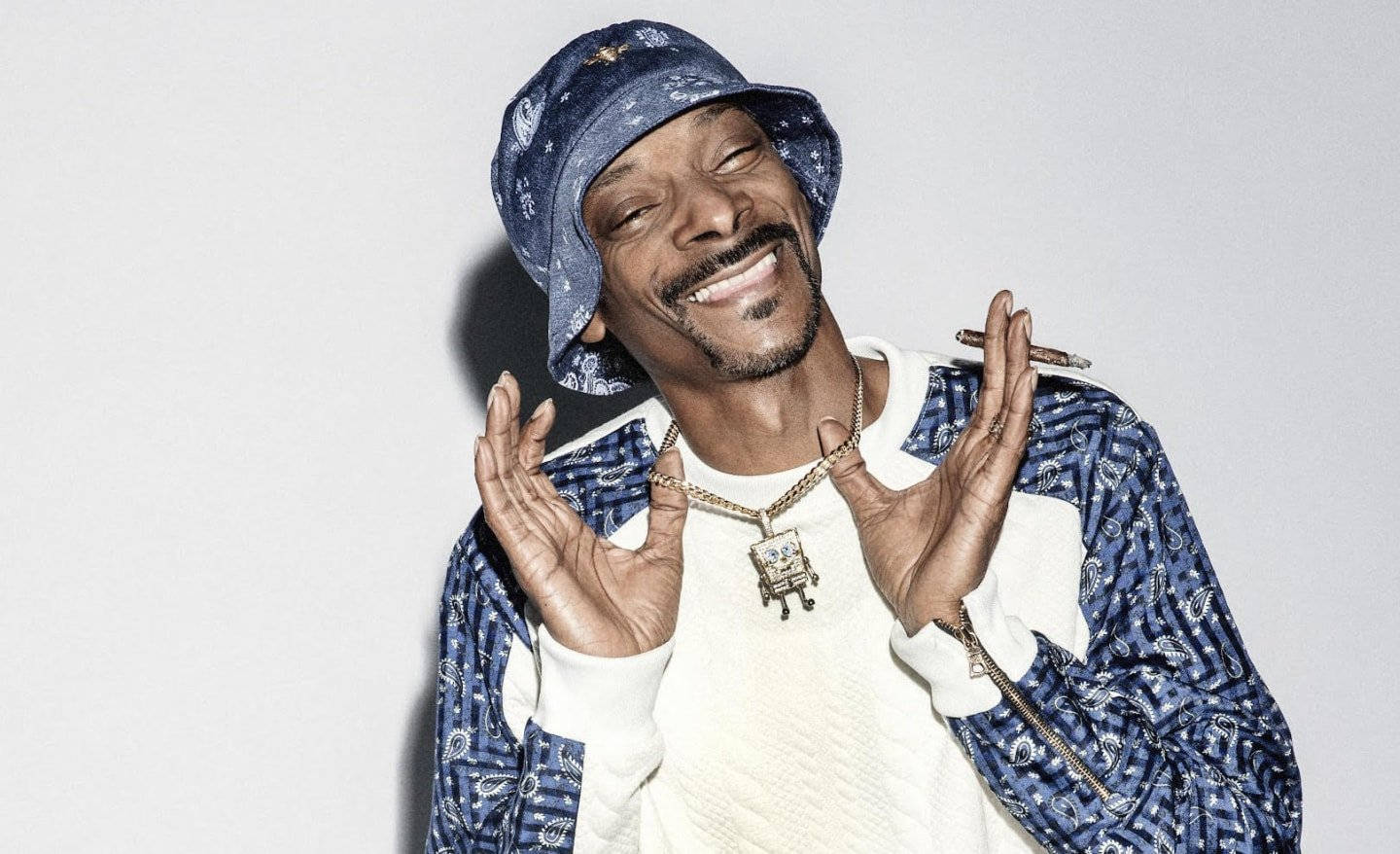 90年代饒舌歌手 桌布 壁紙 — Snoop Dogg 咧嘴大笑，戴著藍色圖案漁夫帽和相配的夾克，背景是純白色。
