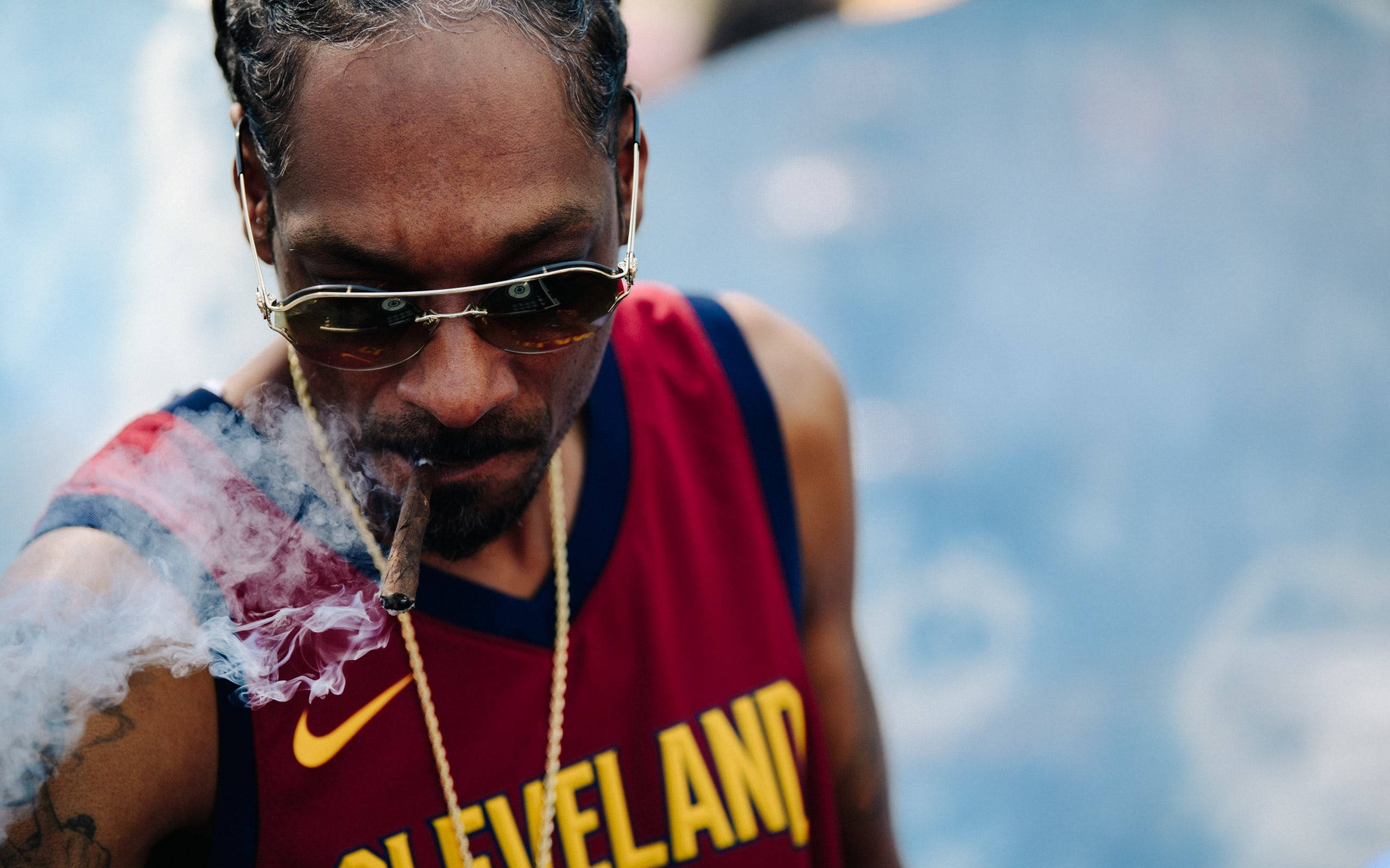 Snoop 桌布 壁紙 — Snoop Dogg 在模糊的藍色背景下，穿著紅色克里夫蘭球衣抽雪茄，煙霧在他臉龐周圍盤旋。