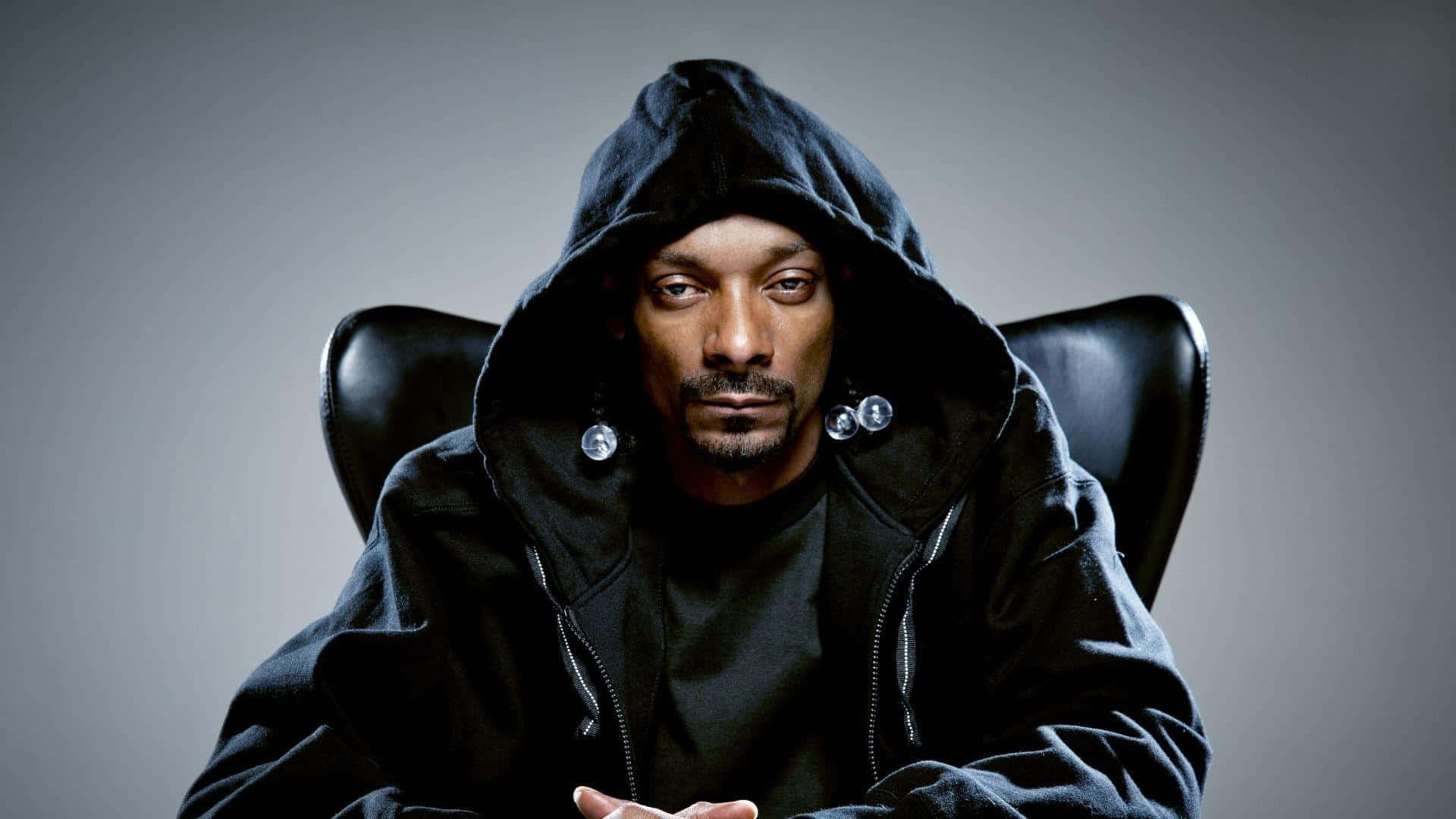 Young Snoop Dogg 桌布 壁紙 — Snoop Dogg 坐在黑色椅子上，身穿黑色連帽衫，戴著銀色耳環，背景為灰色。