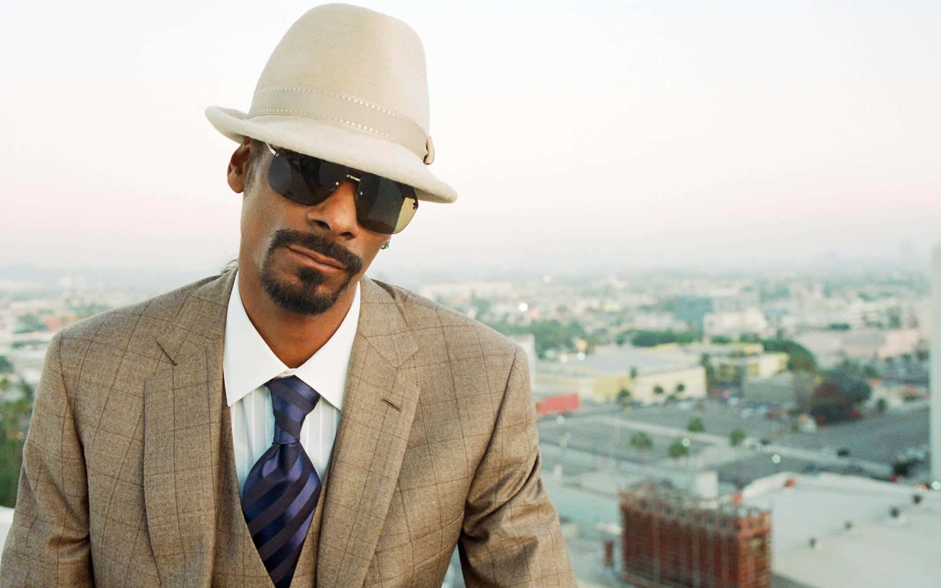 Young Snoop Dogg 桌布 壁紙 — Snoop Dogg 戴著米色紳士帽和太陽眼鏡，在黎明時分的城市天際線上自信地擺姿勢。