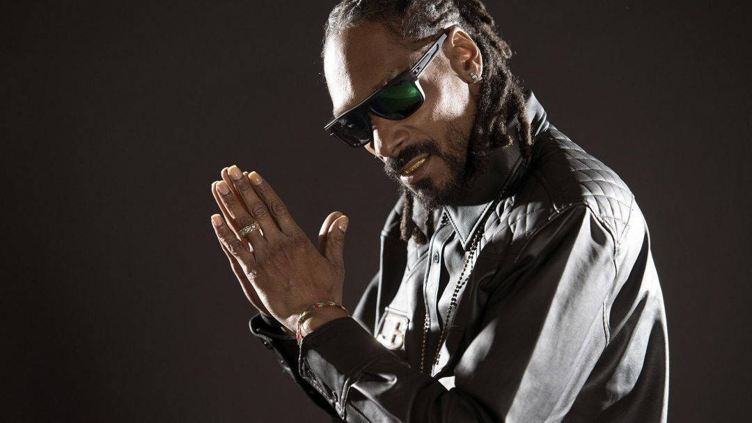 Snoop Dogg 桌布 壁紙 — Snoop Dogg 戴著黑色太陽眼鏡和外套，在昏暗的攝影棚肖像中雙手合十，燈光效果戲劇化。