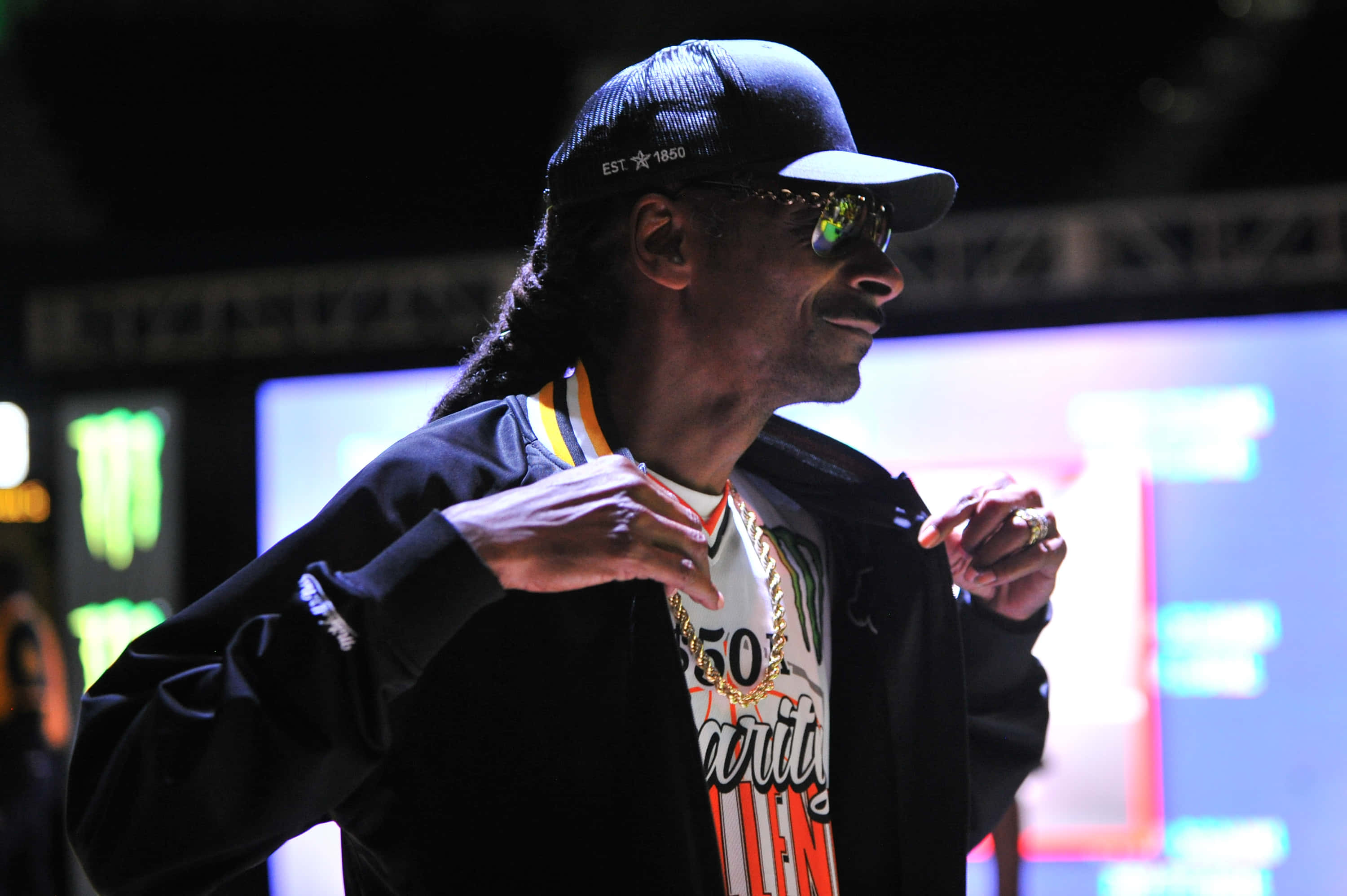 Young Snoop Dogg 桌布 壁紙 — Snoop Dogg 戴著黑色帽子和外套，在明亮的舞台燈光下自信地比手勢，身後有發光的 Monster Energy 標誌。