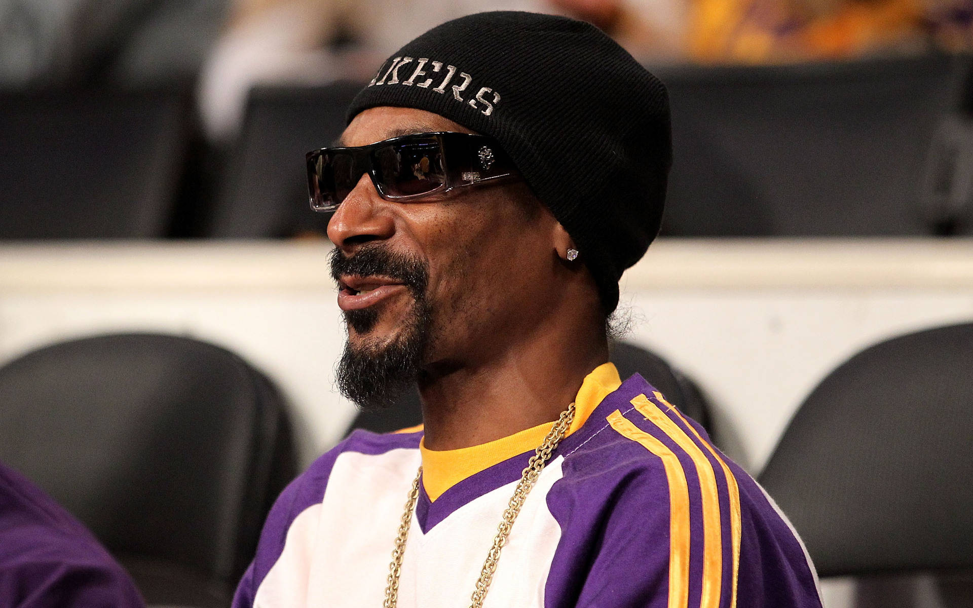 Snoop 桌布 壁紙 — Snoop Dogg 戴著黑色毛帽和太陽眼鏡，身穿紫色和金色的湖人球衣坐在體育館座位上。