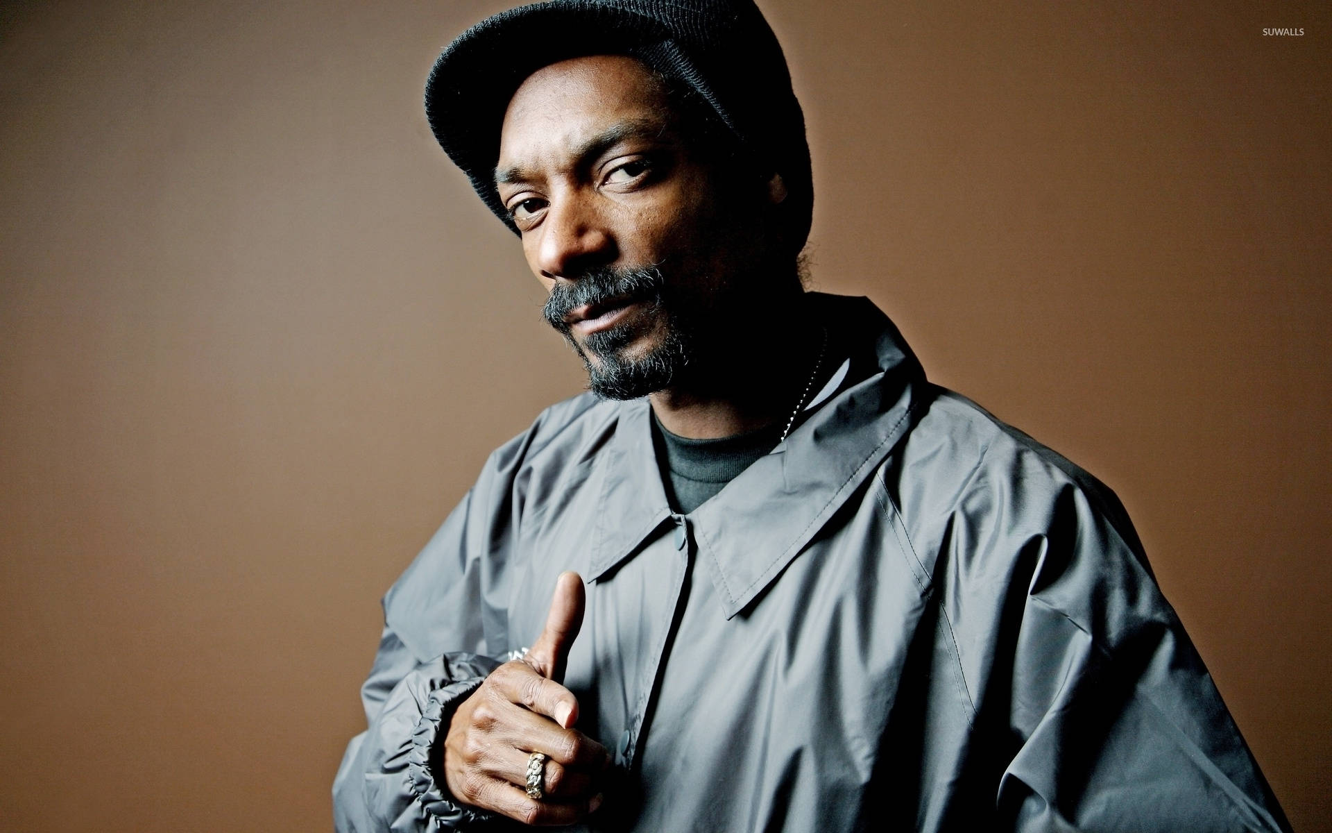 Snoop 桌布 壁紙 — Snoop Dogg 穿著灰色外套和黑色毛帽，在溫暖的棕色背景下自信地豎起大拇指。