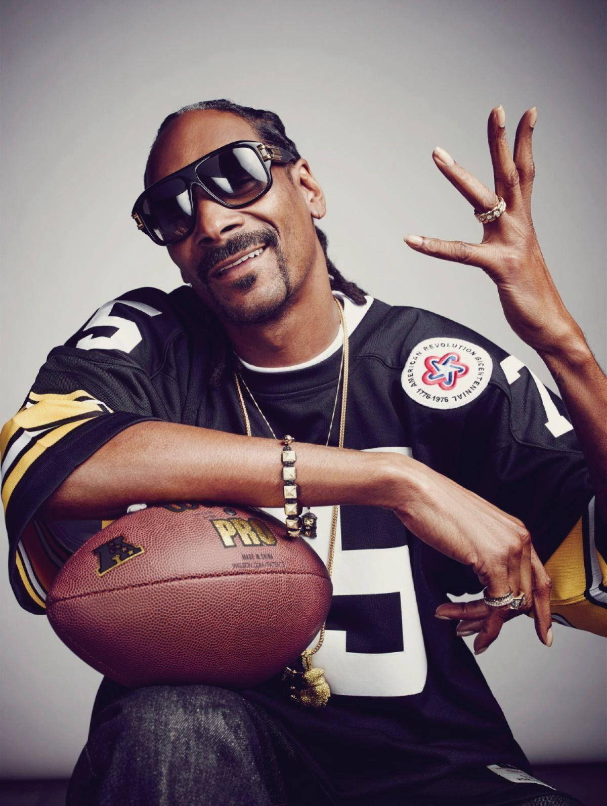 Snoop 桌布 壁紙 — Snoop Dogg 自信地微笑，身穿黑金色美式足球球衣，手持棕色皮革美式足球，背景為純灰色。