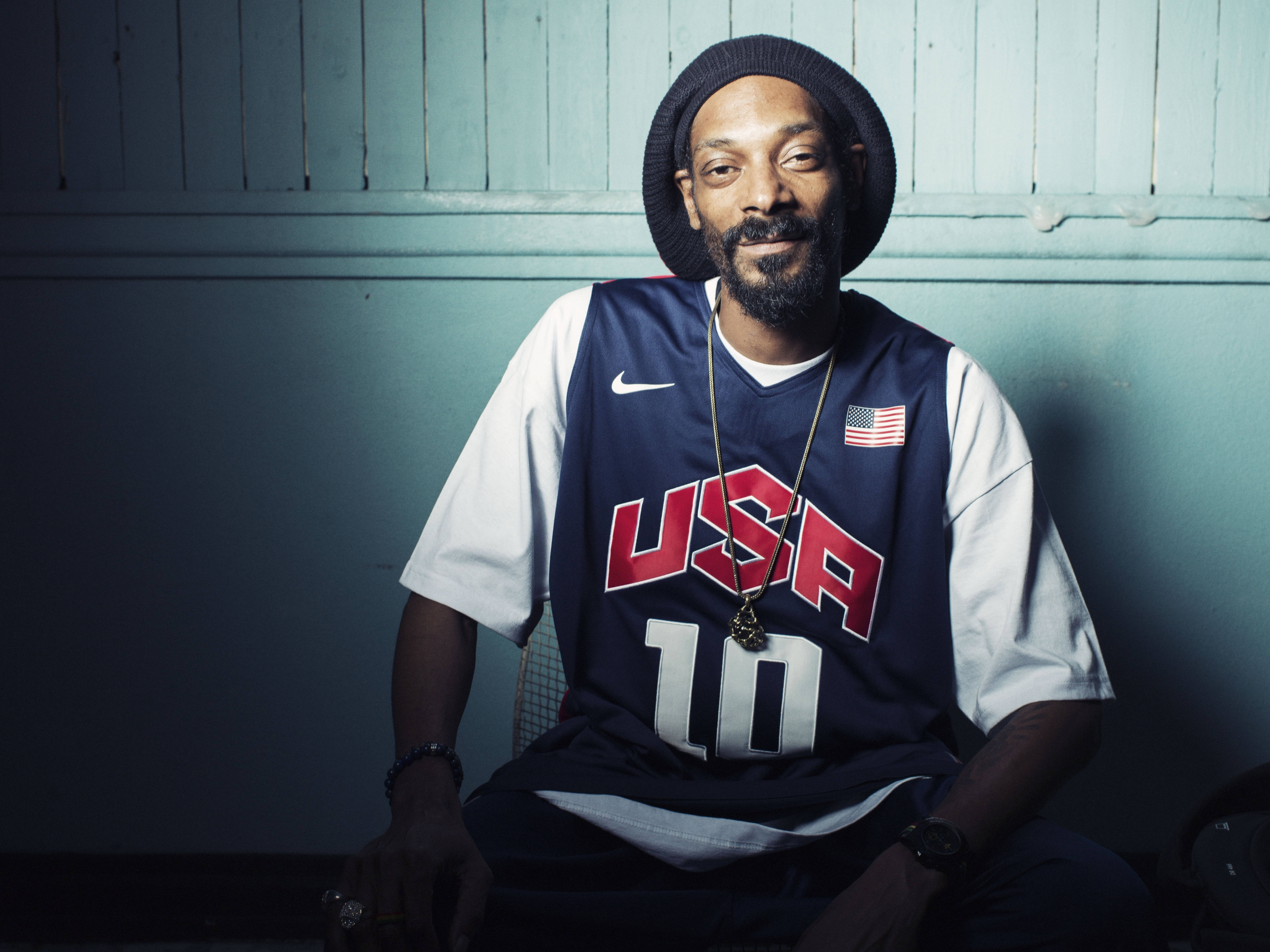Snoop 桌布 壁紙 — Snoop Dogg 隨意地坐在燈光昏暗的房間裡，身穿美國籃球球衣和毛帽，散發著酷酷的自信。