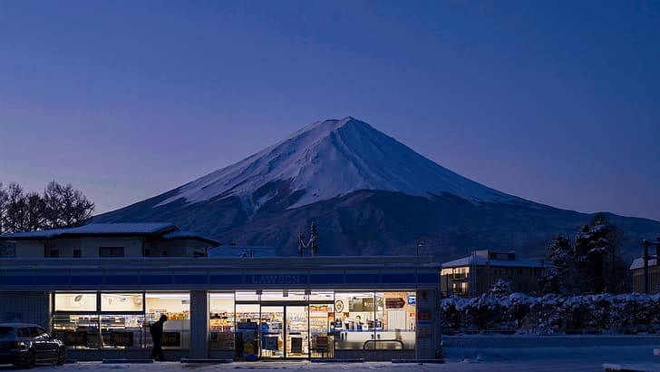 白雪覆蓋的富士山在暮色中聳立在發光的Lawson店後面。 — 4K 壁紙