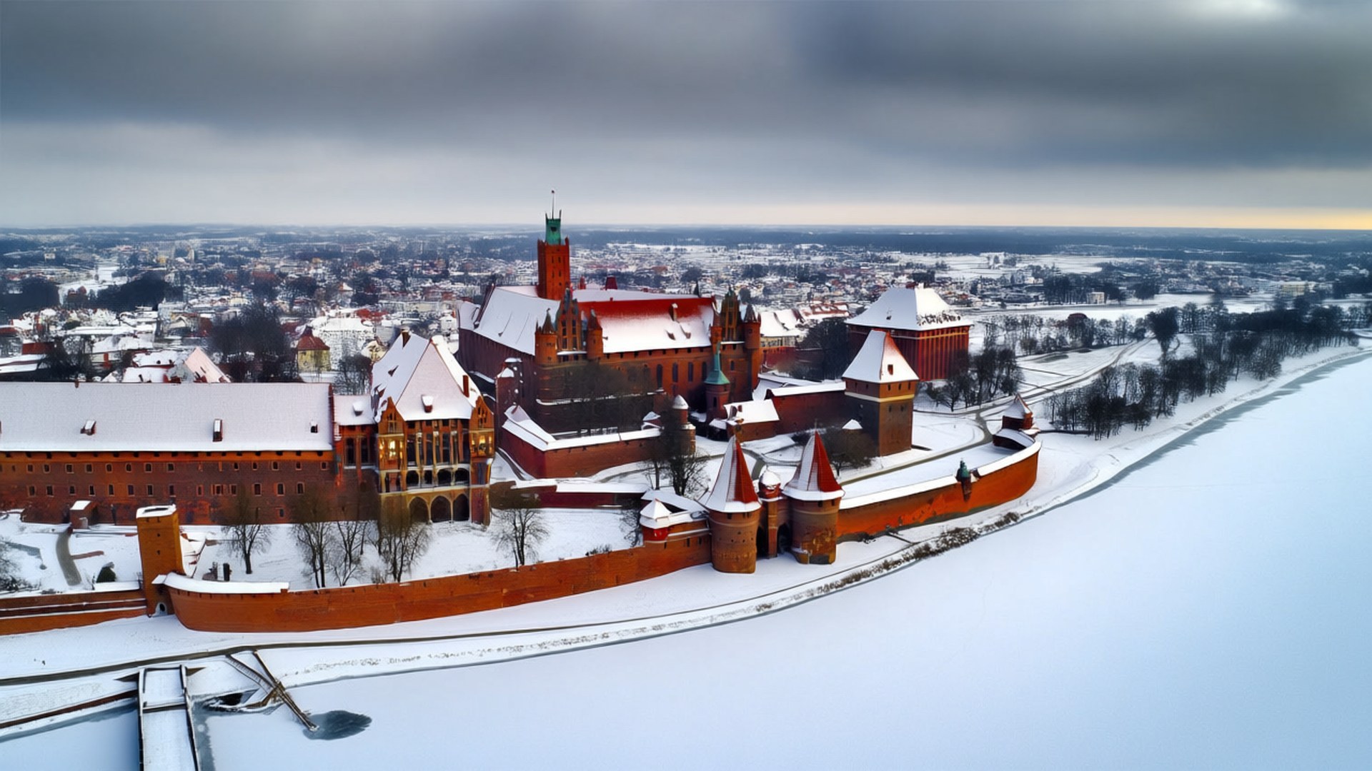 Malbork Castle Winter 壁紙 — 一個被雪覆蓋的紅磚城堡建築群，帶有塔樓和角樓，俯瞰著結冰的河流和城市，天空多雲。