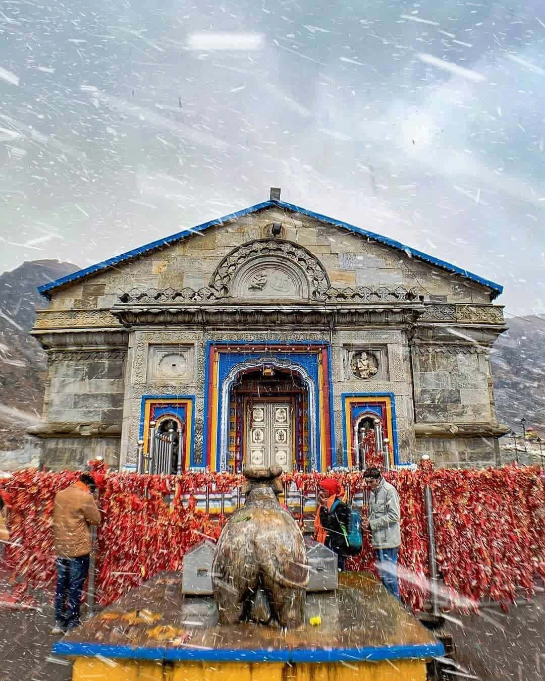 Kedarnath 桌布 壁紙 — 大雪圍繞著一座宏偉的藍邊石廟飄落，人們站在紅色經幡圍欄和公牛雕像附近。