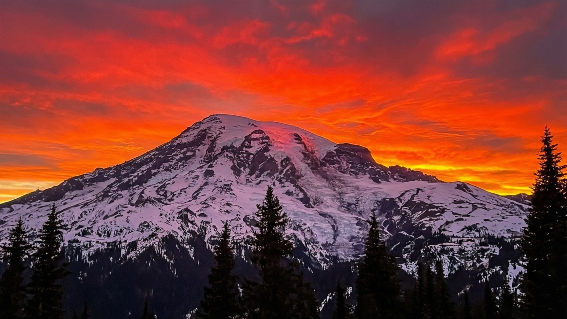 Fiery Sunset Snow 壁紙 — 一座積雪的山峰在火紅橙色的日落天空下升起，前景有常綠樹的剪影。
