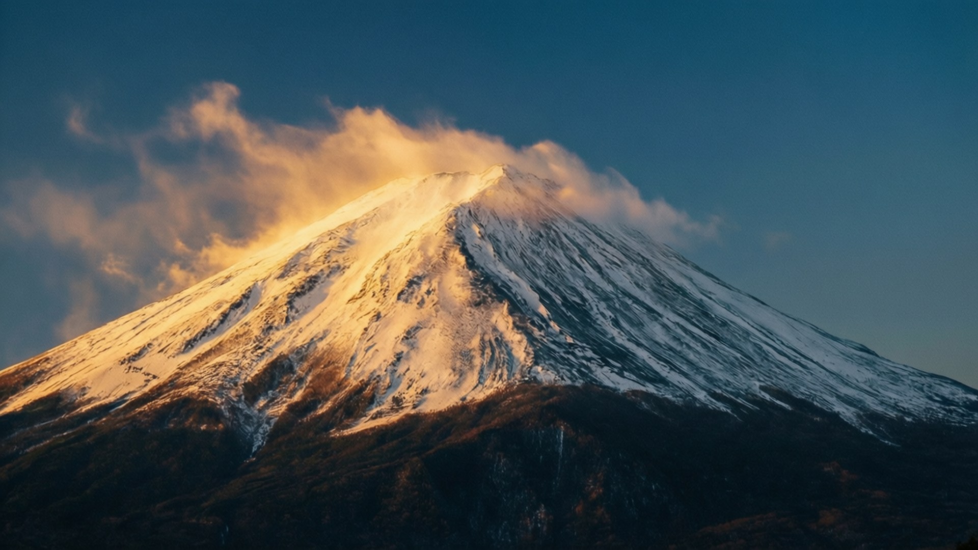 Golden Peak Dawn 壁紙 — 白雪覆蓋的山峰沐浴在金色的日出光芒中，縷縷雲彩在其山頂盤旋，映襯著深藍色的天空。
