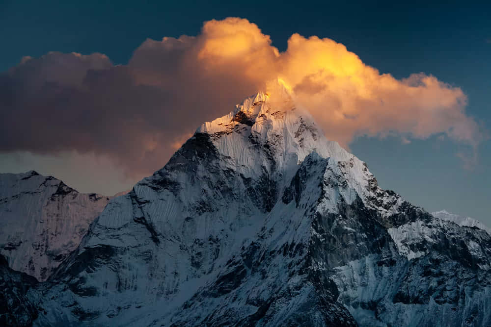  壁紙 — 白雪覆蓋的山峰刺穿戲劇性的雲層，在日落時分映襯著深藍色的天空閃耀金光。