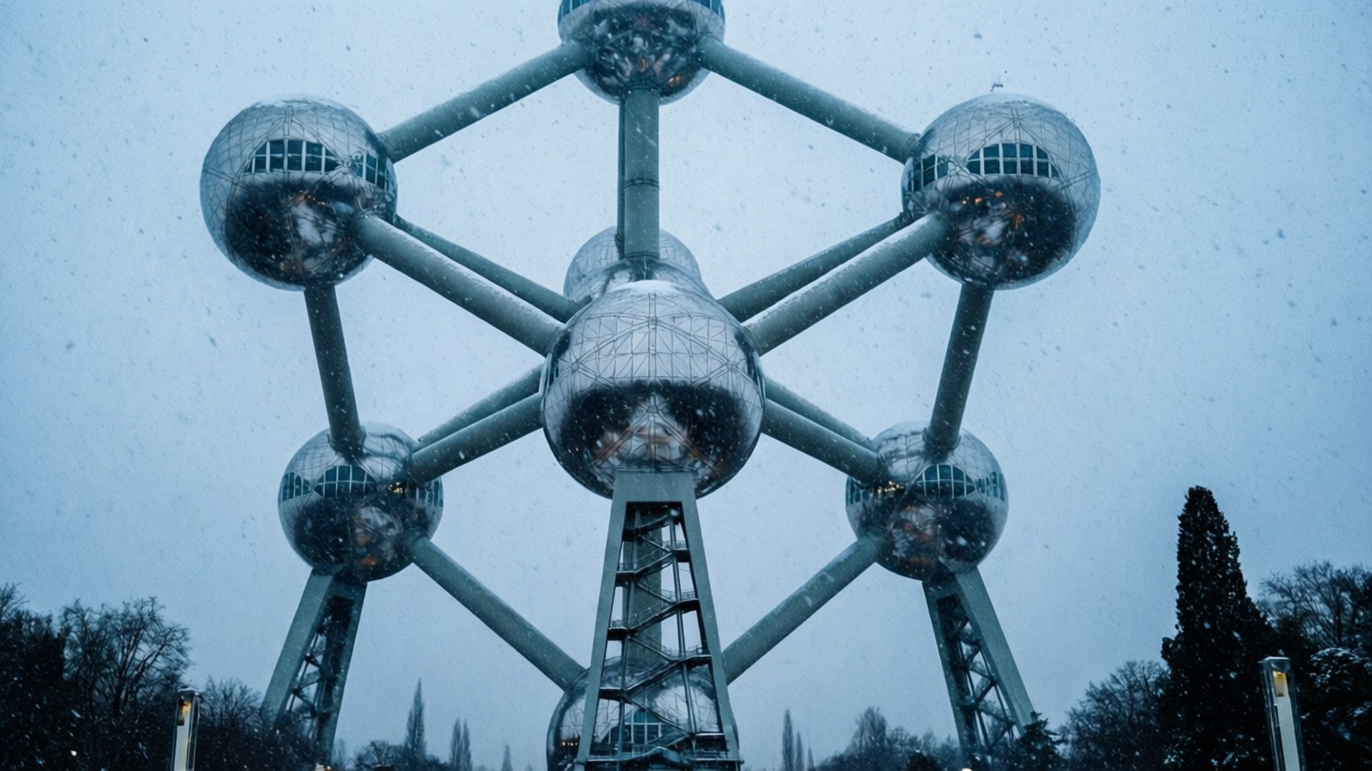 Snowy Atomium 壁紙 — 雪花在灰色冬日天空下飄過標誌性的原子球塔結構，背景是光禿禿的樹木。