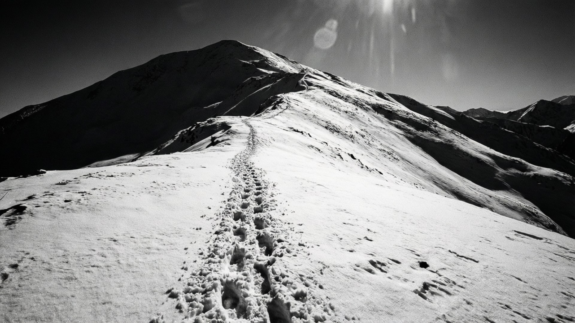 Footprints in Snow 壁紙 — 一條孤獨的腳印蜿蜒向上，穿過純淨的雪地，通往一個陽光照耀的山峰，天空呈現鮮明的黑白對比。