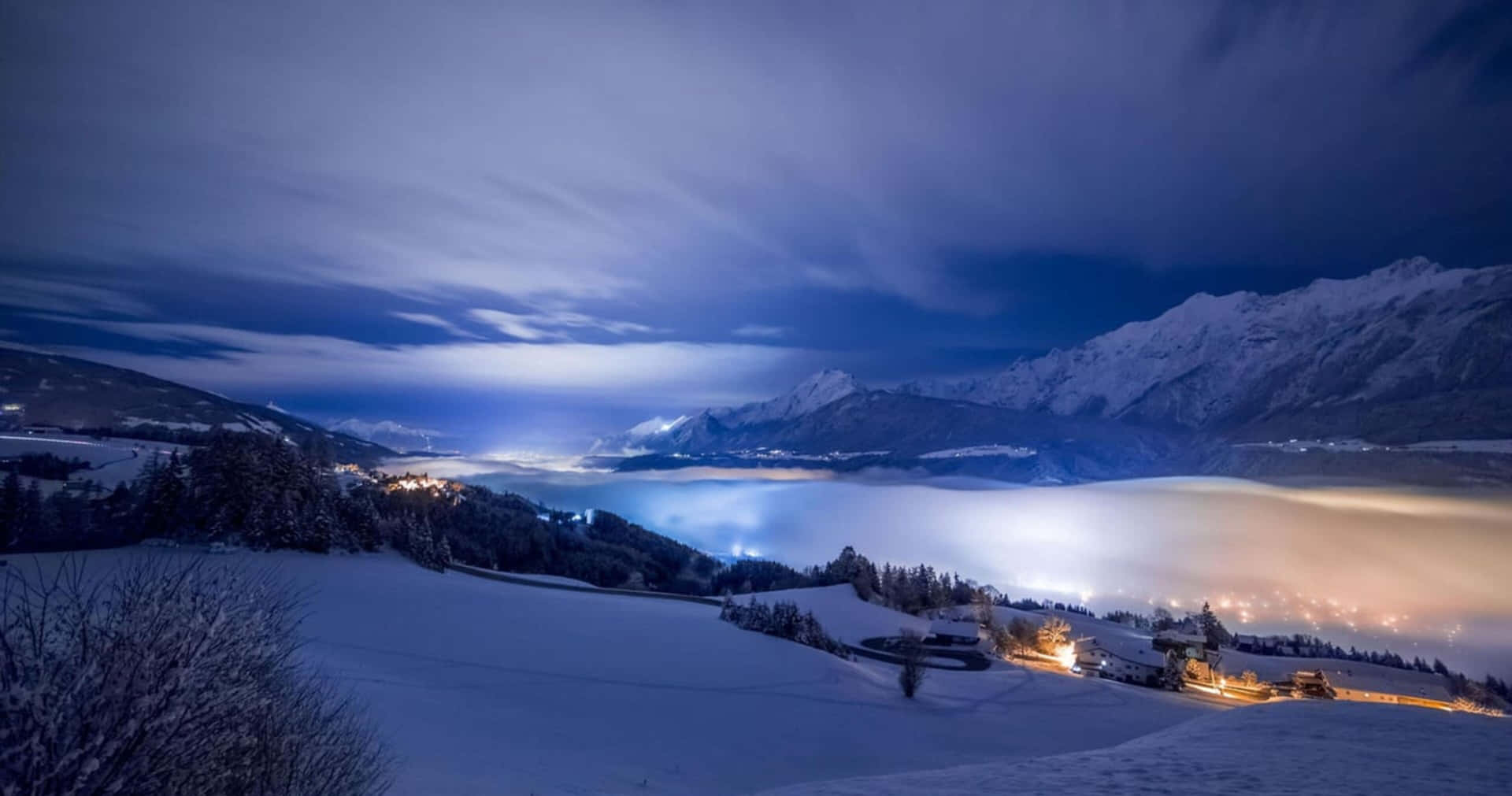 夜晚白雪覆蓋的山谷，村莊燈光在黑暗雲層下閃爍。 — 4K 壁紙