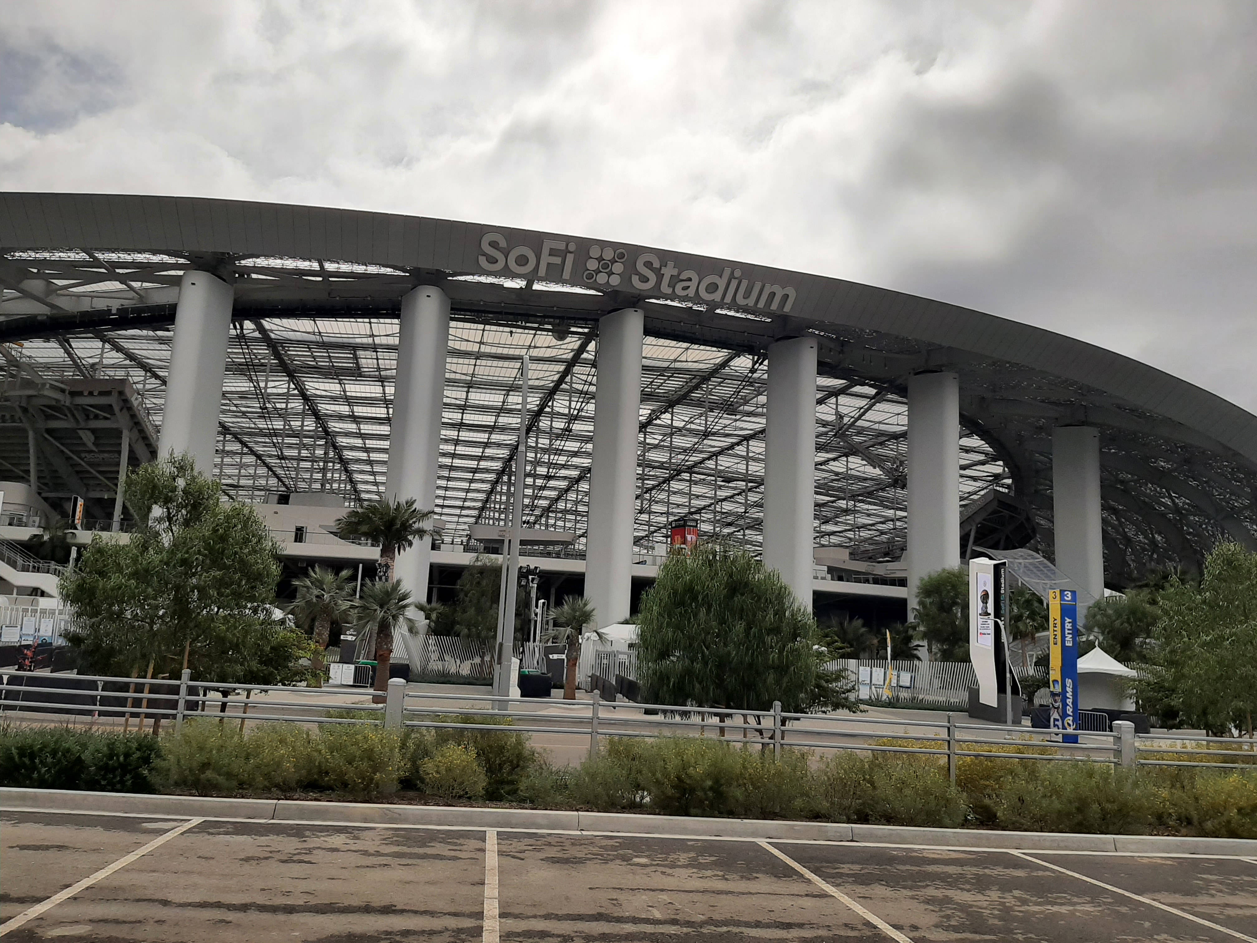 NFL 美式足球 桌布 壁紙 — Sofi Stadium 矗立於多雲的天空下，前景有棕櫚樹和停車場。