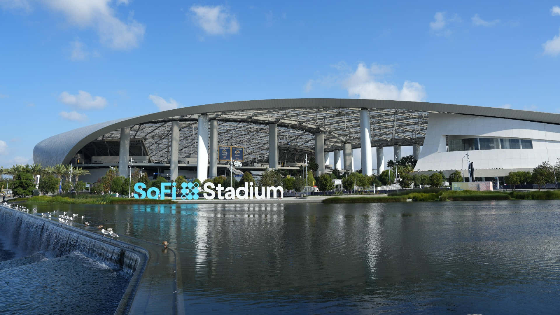 Sofi Stadium 桌布 壁紙 — SoFi 體育場在明亮的藍天白雲下閃耀，其現代建築倒映在平靜的水面上，鳥兒聚集在小瀑布附近。