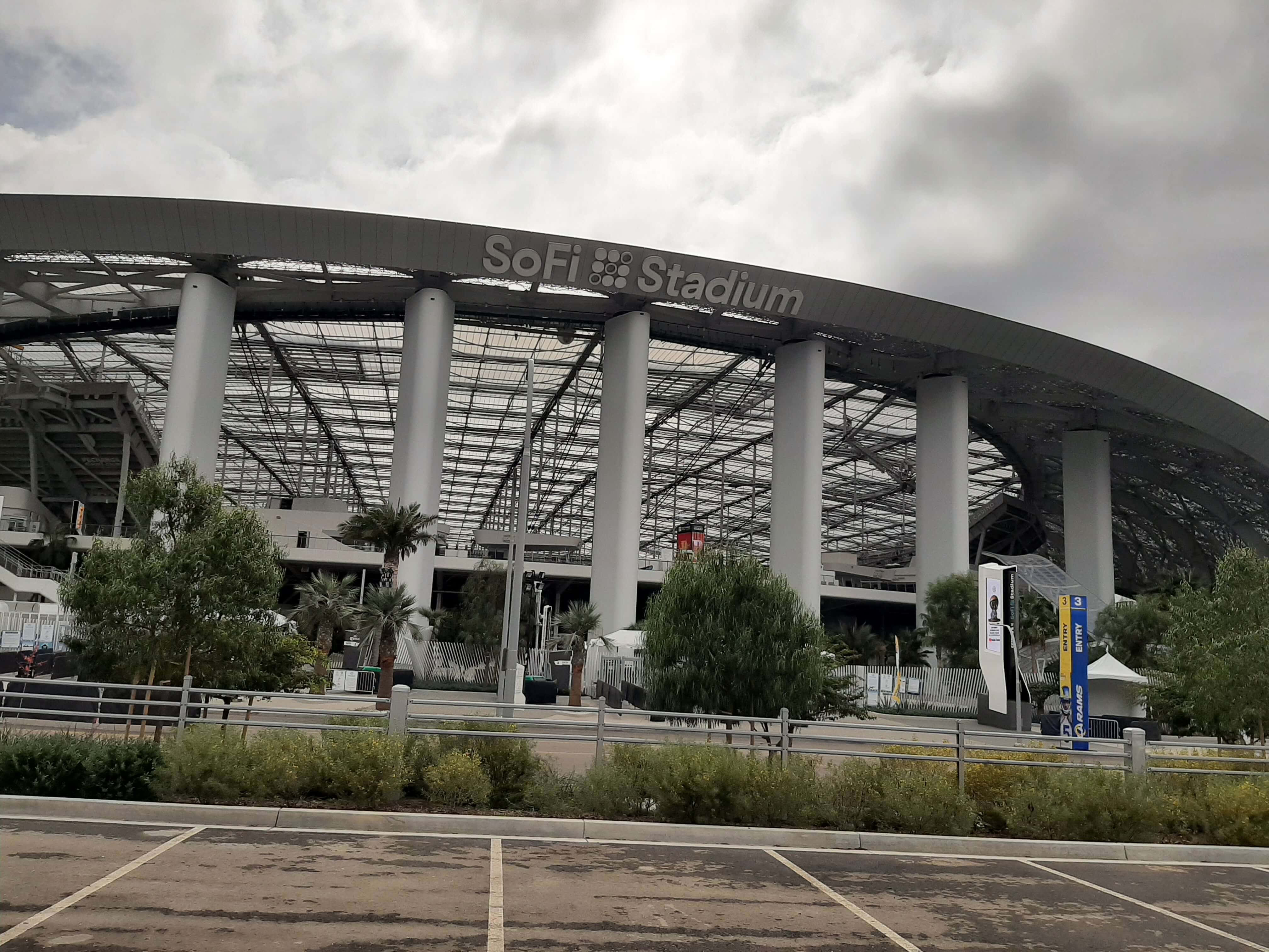 Sofi Stadium 桌布 壁紙 — SoFi 體育場在陰天之下，其現代鋼鐵和玻璃結構被綠地和停車場環繞。