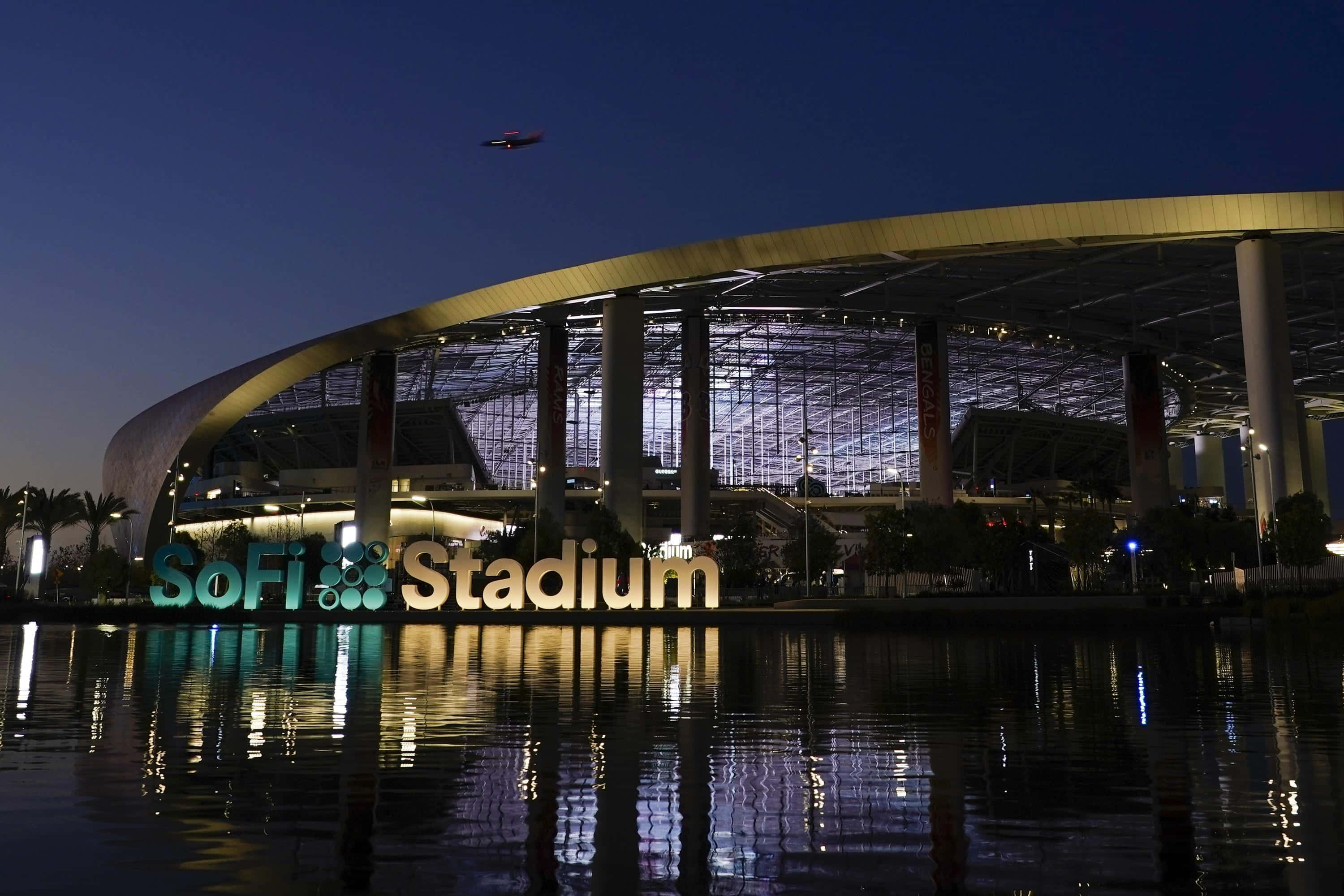 Sofi Stadium 桌布 壁紙 — SoFi 體育場在黃昏時分閃耀，其發光的標誌倒映在平靜的水面上，一架無人機在上空飛過。