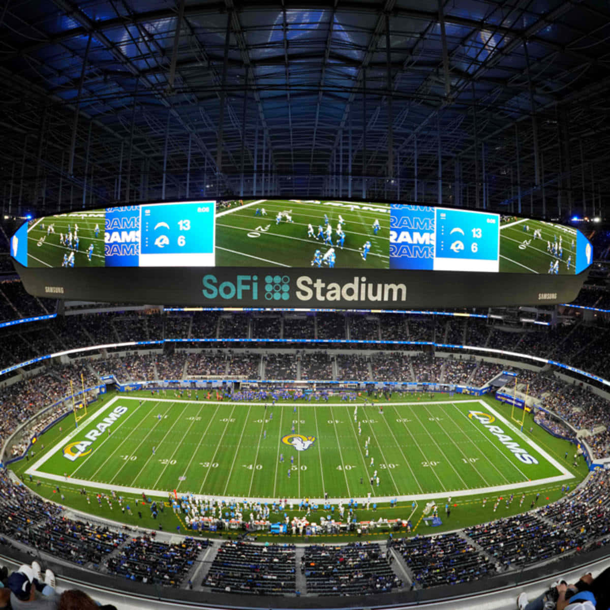 Sofi Stadium 桌布 壁紙 — SoFi 體育場夜景鳥瞰圖，藍色燈光閃爍，照亮了公羊隊比賽期間擠滿觀眾的看台和充滿活力的綠色球場。