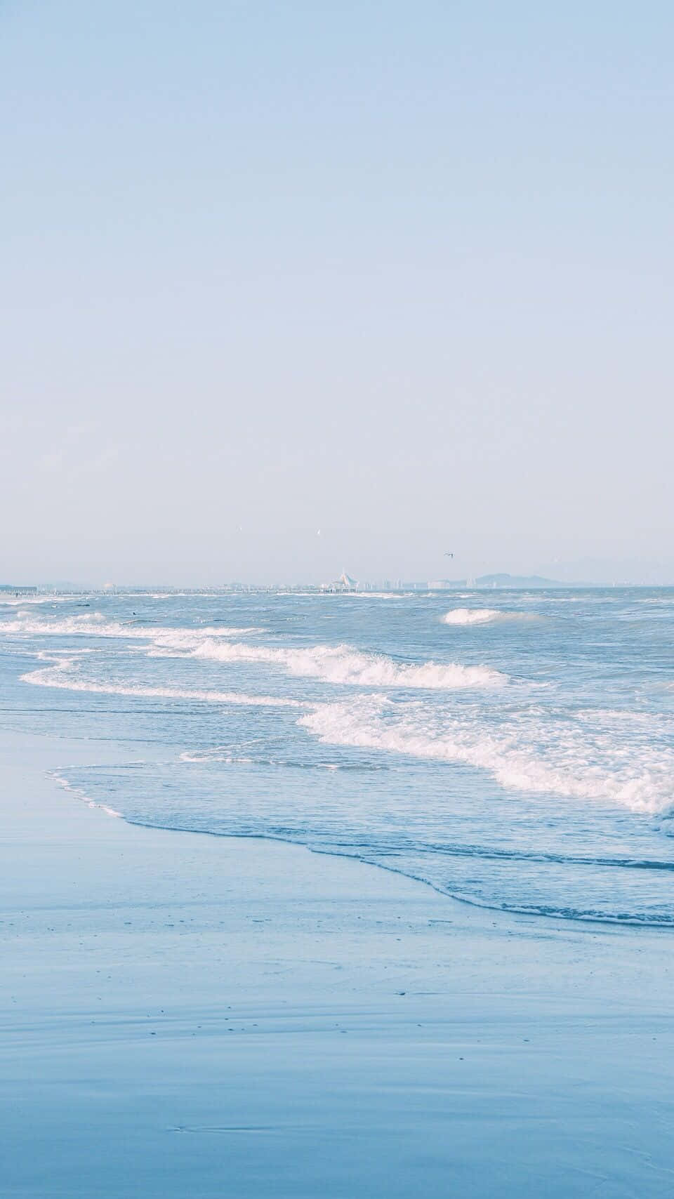  壁紙 — 柔和的藍色海浪輕輕地拍打著寧靜的海灘，天空清澈蒼白，地平線上有遙遠的陸地。