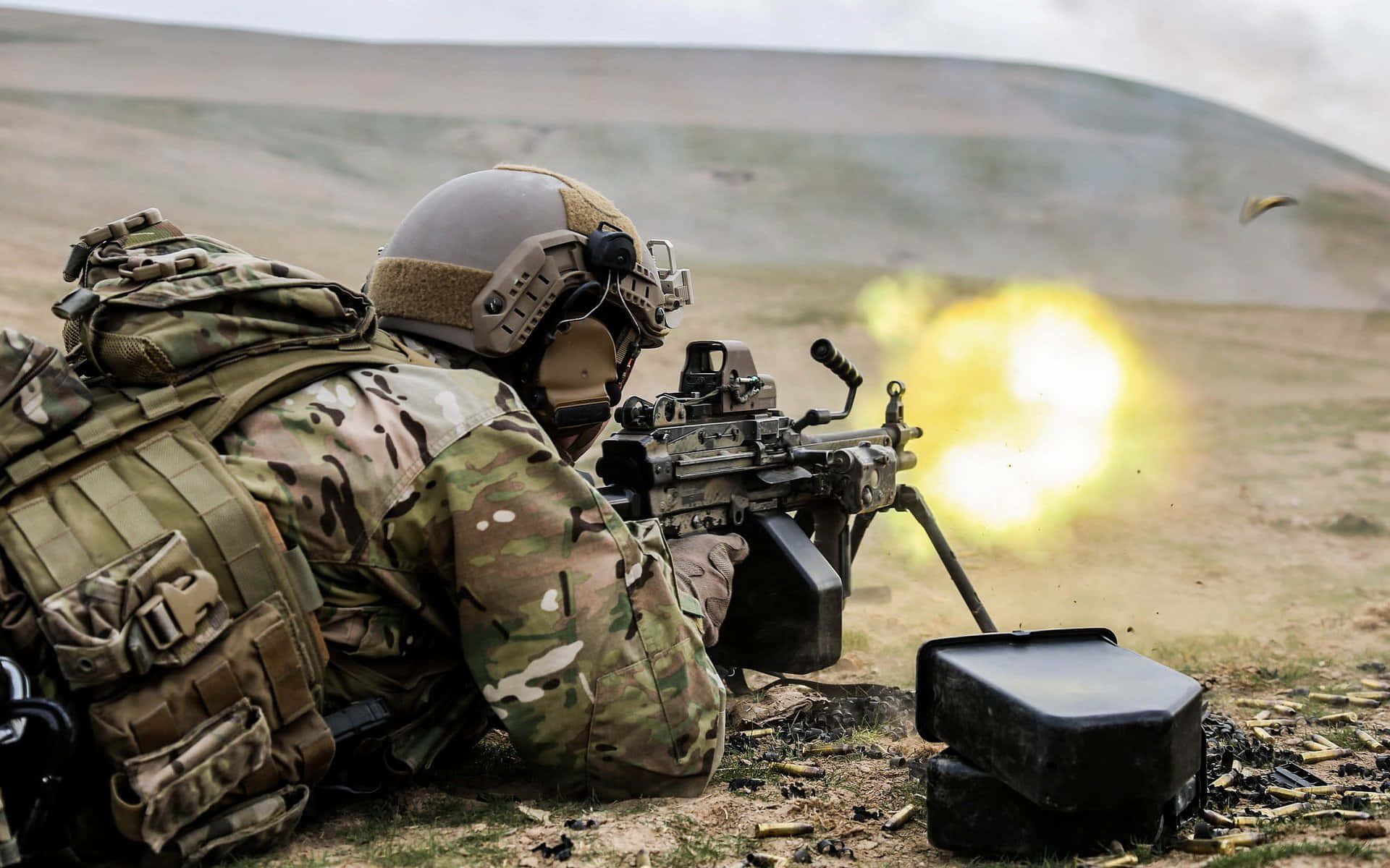  壁紙 — 一名身穿迷彩服的士兵在荒蕪的田野中發射重型機槍，伴隨著明亮的槍口閃光及散落的彈殼。