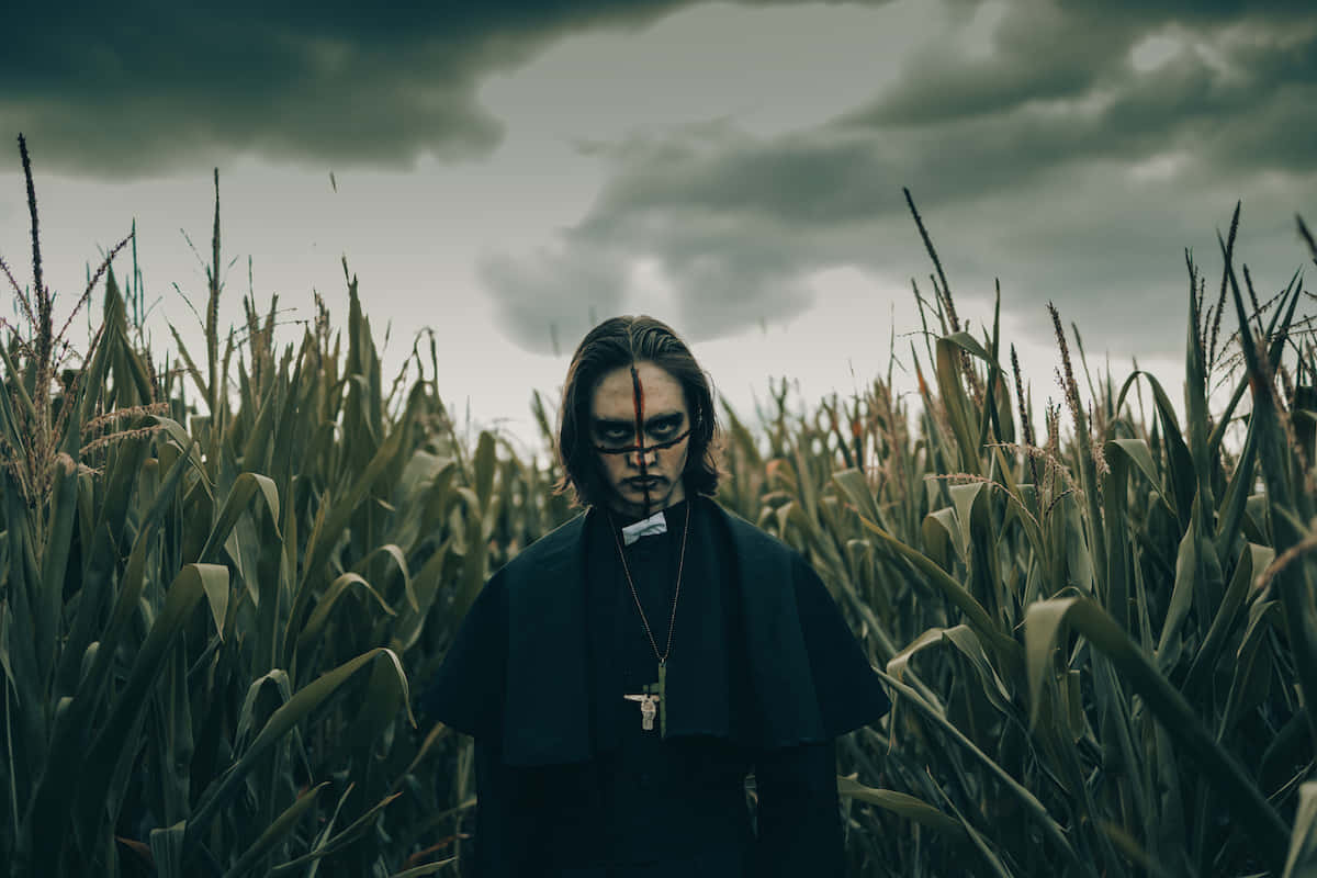  壁紙 — 一個身穿黑色神職服裝的莊嚴人物，矗立在陰沉多雲的天空下高聳的玉米田中。