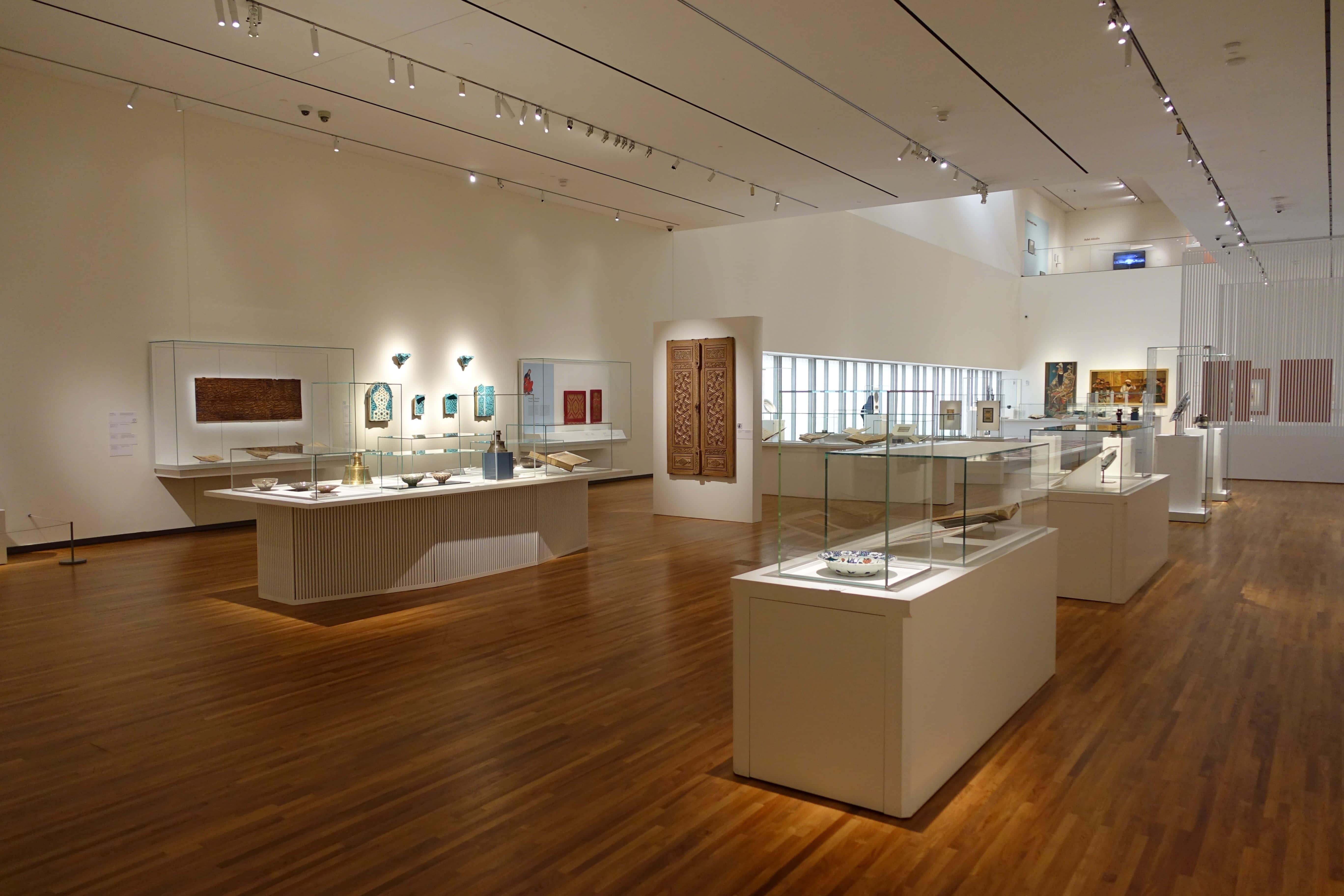 Aga Khan Museum 桌布 壁紙 — 寬敞的博物館畫廊，拋光木地板和白色展示櫃在聚焦燈光下展示古代文物。