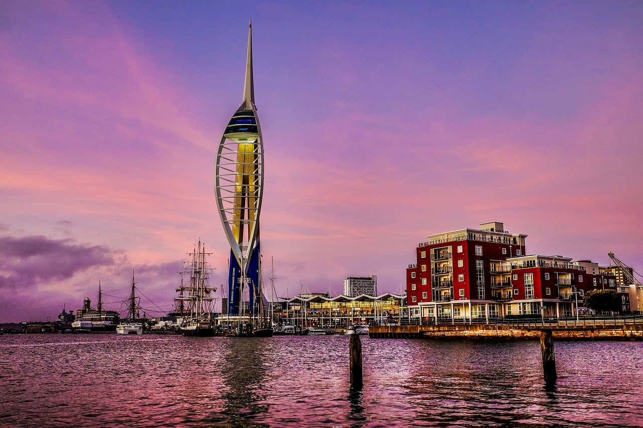 UK 桌布 壁紙 — Spinnaker Tower 在充滿活力的粉紅色和紫色日落天空下高聳，其燈光倒映在平靜的港灣水面上。