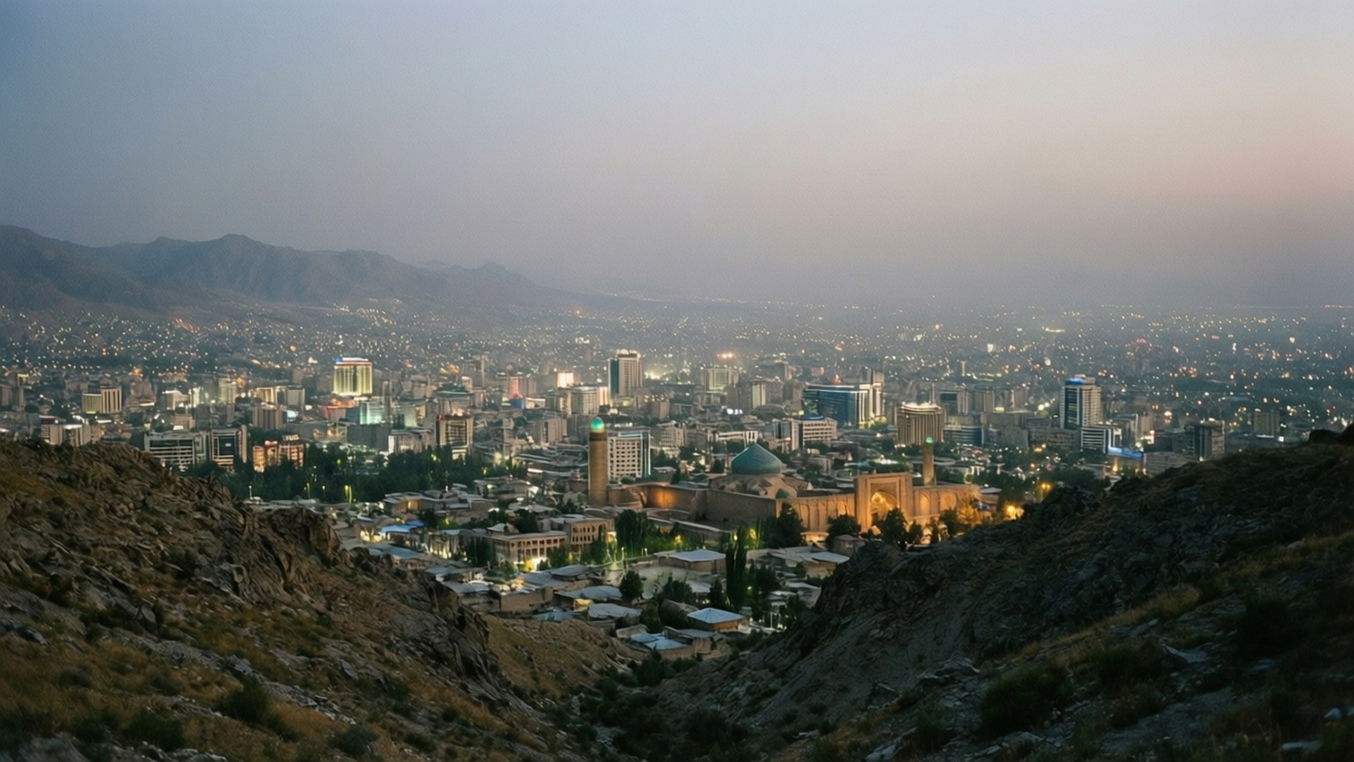Kabul City Lights 壁紙 — 在柔和的漸變天空下，廣闊的城市景觀在黃昏時分，發光的建築物坐落在崎嶇的山脈之間。