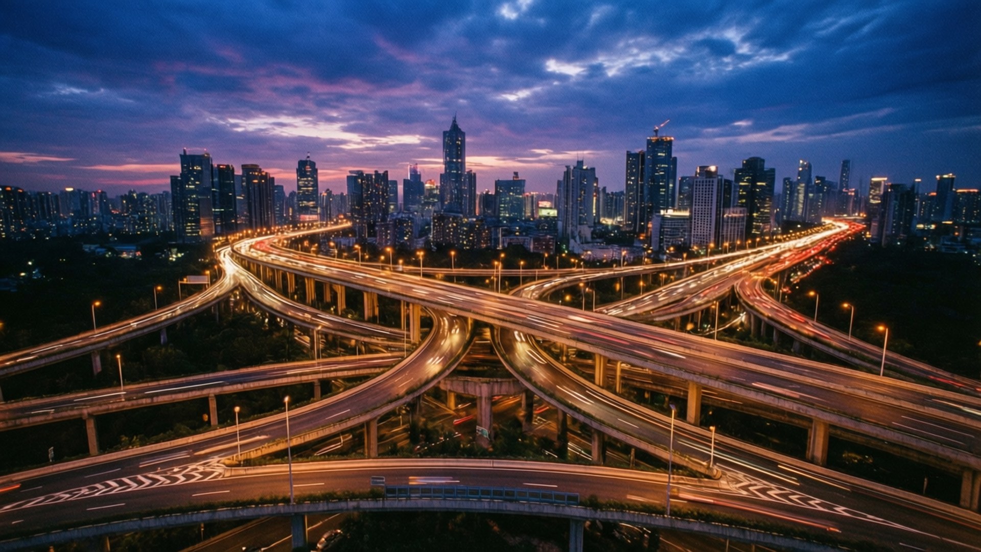 Highway Interchange 壁紙 — 黃昏時分，廣闊的高速公路交匯處，在充滿活力的紫色和藍色天空下，密集的城市天際線上蜿蜒著發光的交通軌跡。