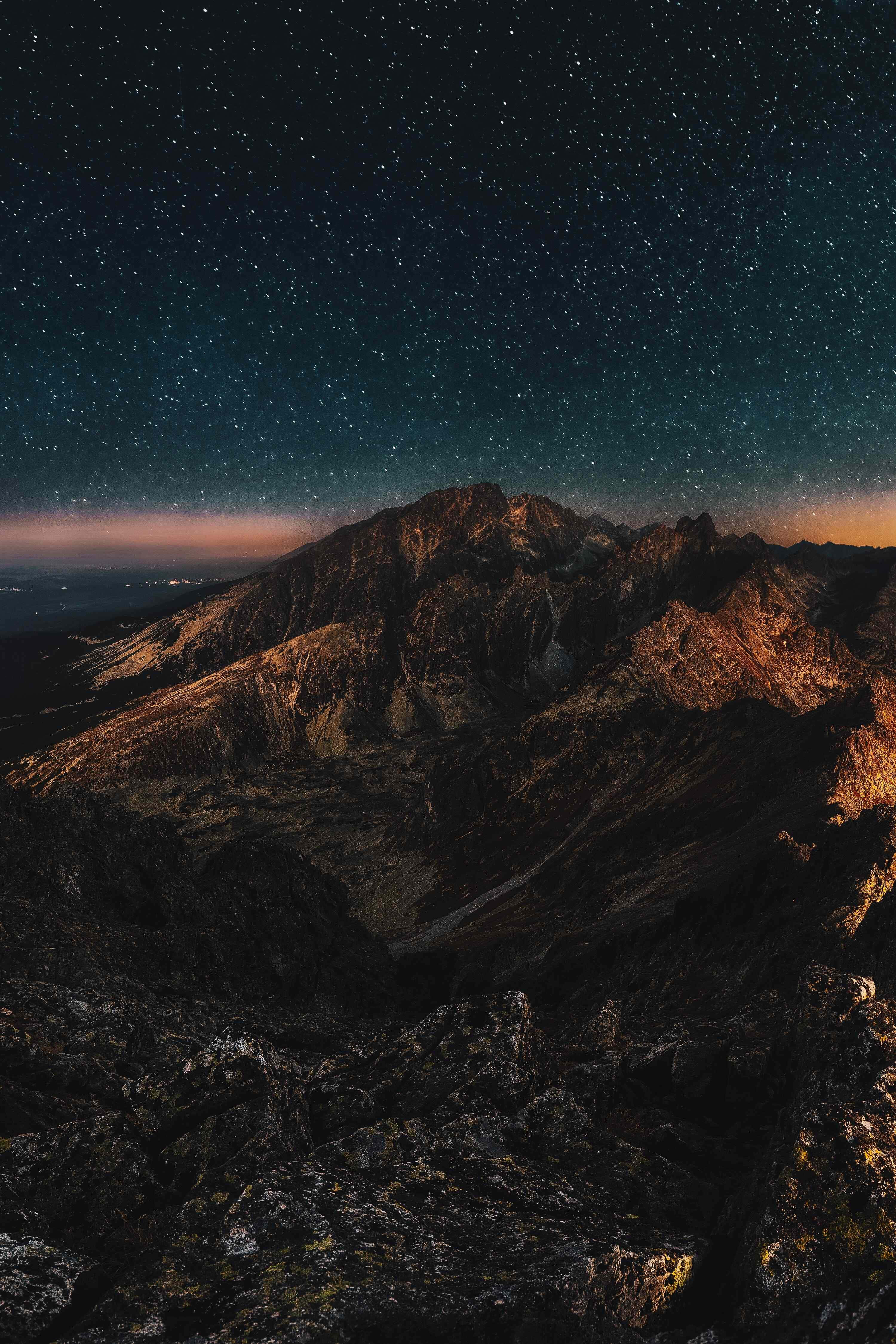 Night Mode 桌布 壁紙 — 繁星點點的夜空在崎嶇的山峰上閃耀，溫暖的橙色光芒照亮了岩石地形。