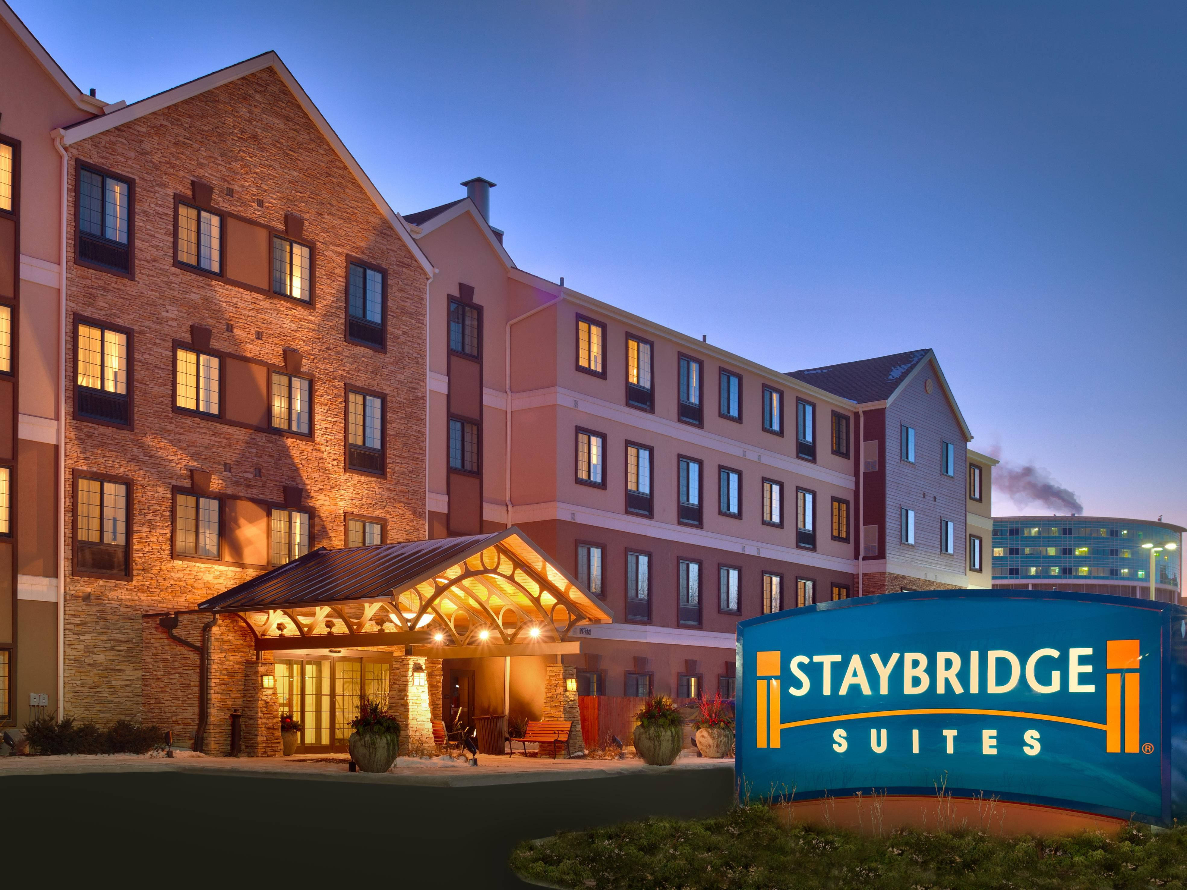 Hotel 桌布 壁紙 — Staybridge Suites 飯店外觀，暮色中溫暖發光的窗戶和顯眼的藍色標誌。