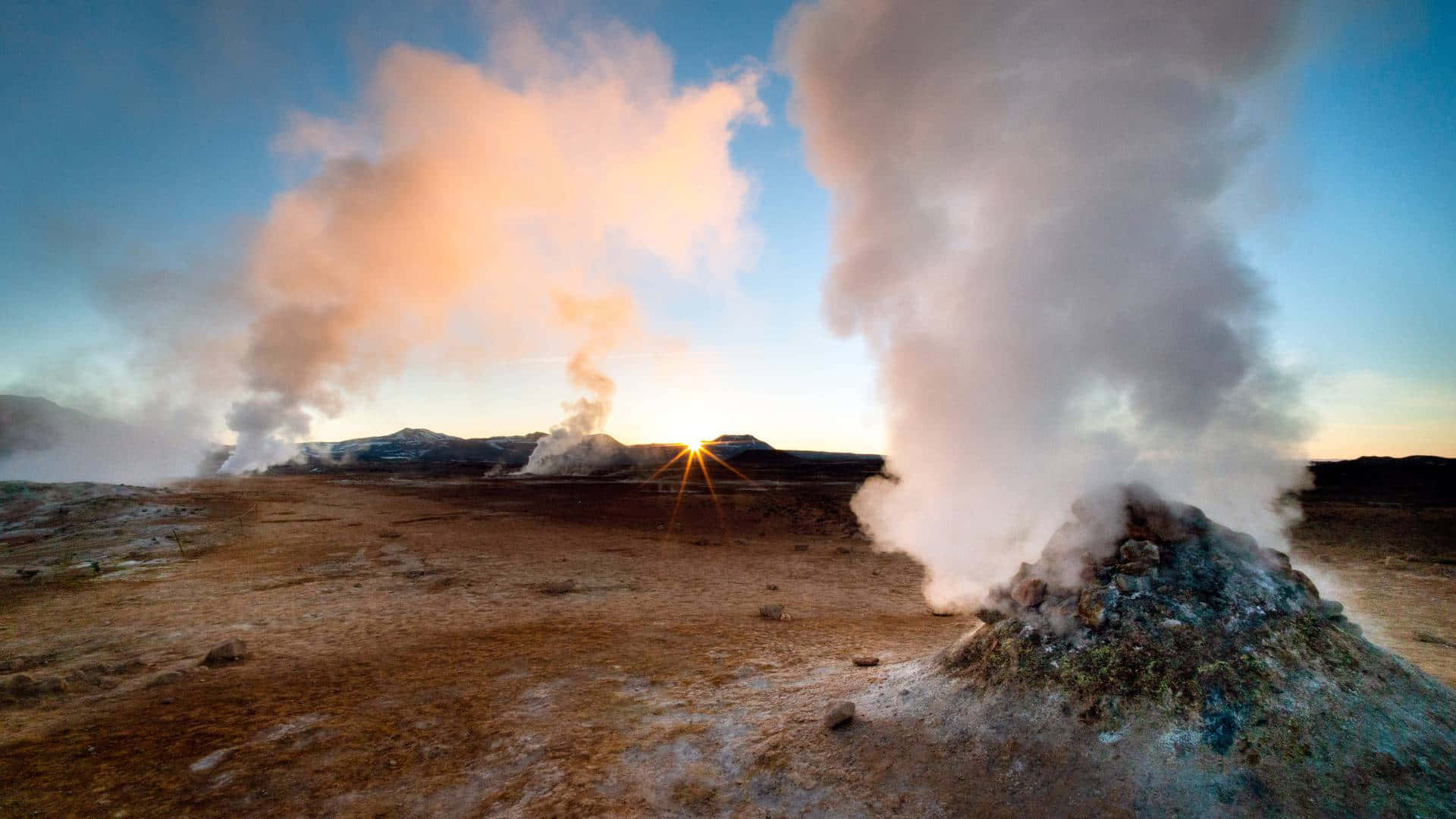 壁紙 — 蒸氣從地熱通風口戲劇性地升起，在荒蕪的景觀中，太陽在遠山後落下，投射出溫暖的光芒。
