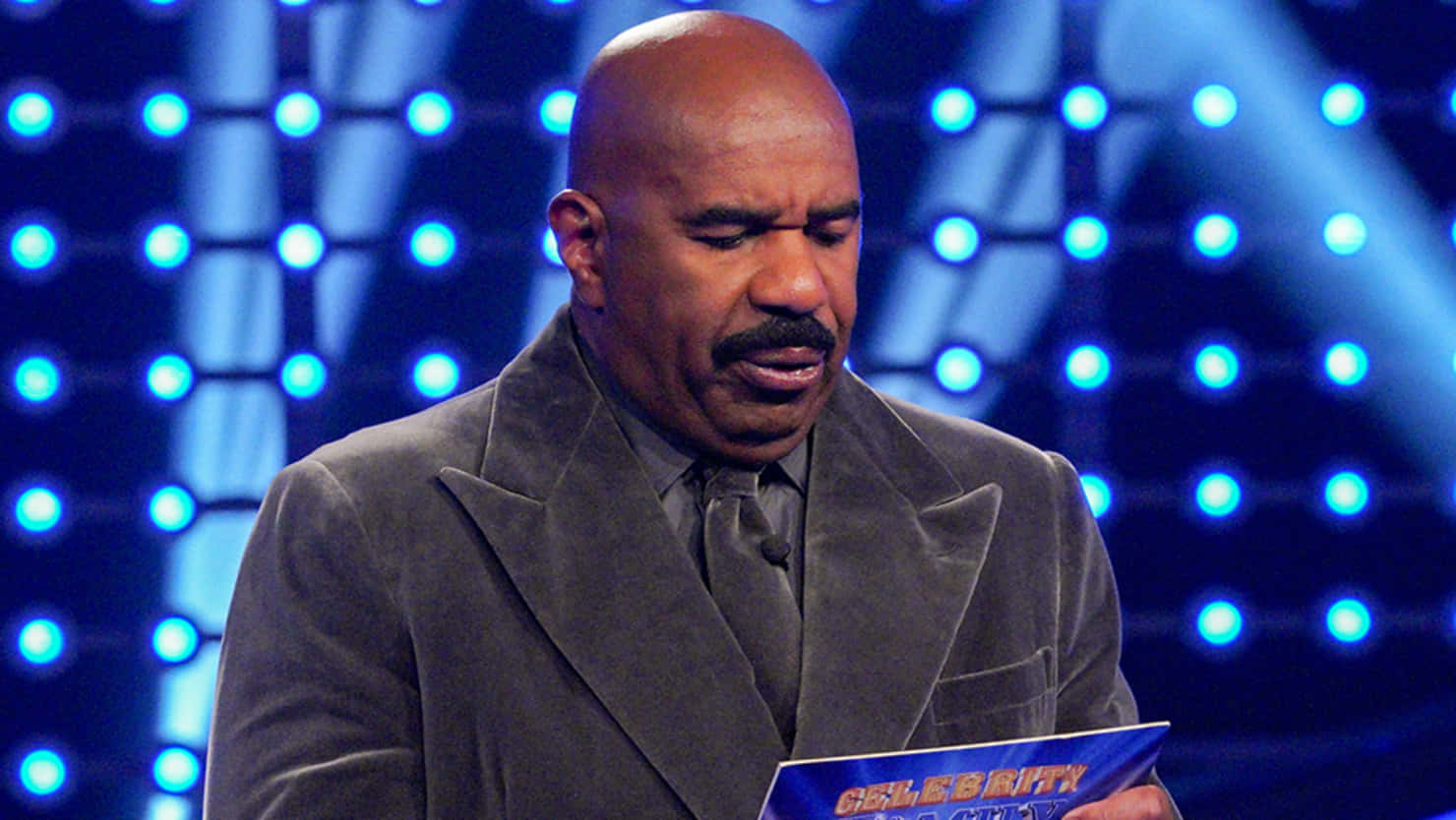 steve harvey 桌布 壁紙 — Steve Harvey 穿著灰色西裝，在發光的藍色舞台燈光背景下拿著一張藍色卡片。