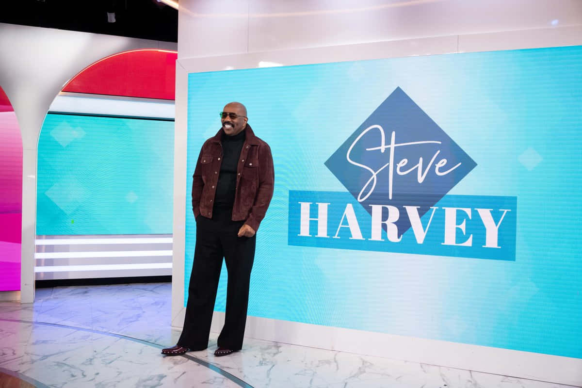 steve harvey 桌布 壁紙 — Steve Harvey 自信地站在一件酒紅色外套前，對著一個顯示他名字的鮮豔藍色螢幕，在現代工作室場景中。