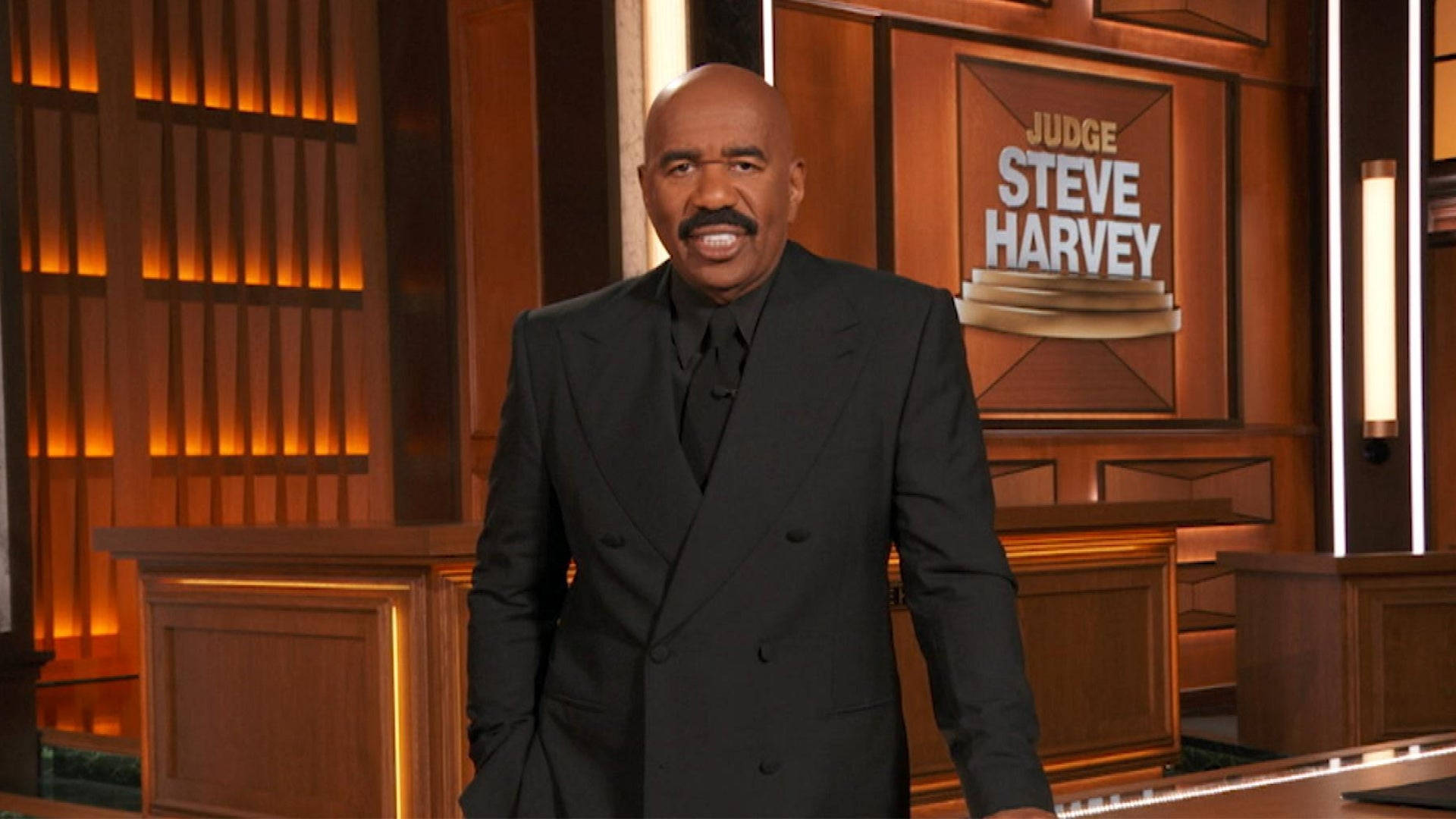 smiling 桌布 壁紙 — Steve Harvey 身穿黑色西裝，自信地站在燈光溫暖的木製舞台上，身後有「Judge Steve Harvey」標誌。