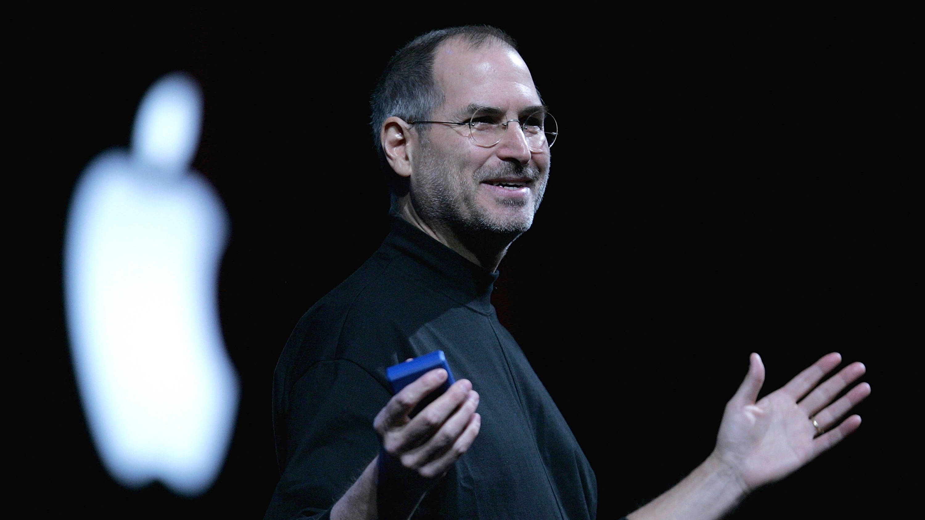 Rich 桌布 壁紙 — Steve Jobs 在舞台上手持藍色裝置做手勢，身後有發光的 Apple 標誌。