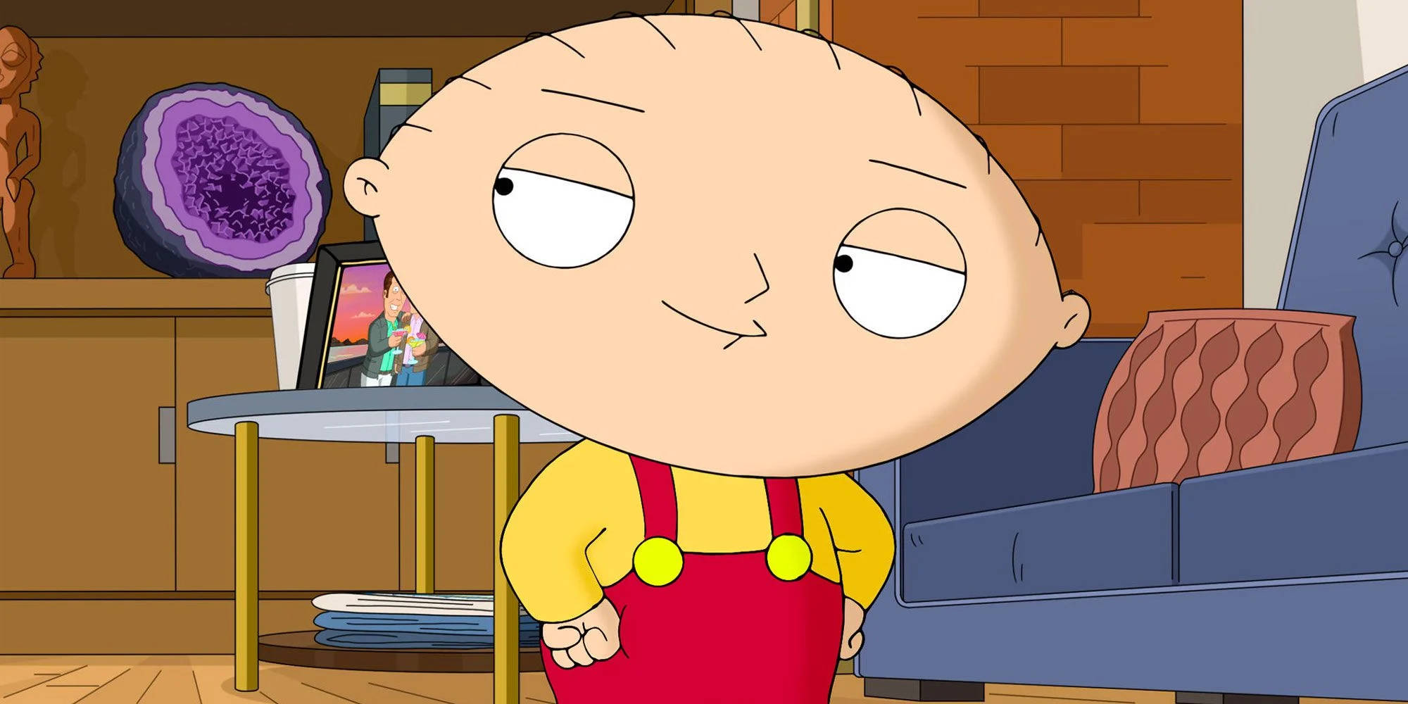 stewie 桌布 壁紙 — Stewie Griffin 身穿黃色襯衫和紅色工裝褲，站在舒適的客廳中，身後有紫色晶洞和藍色沙發。
