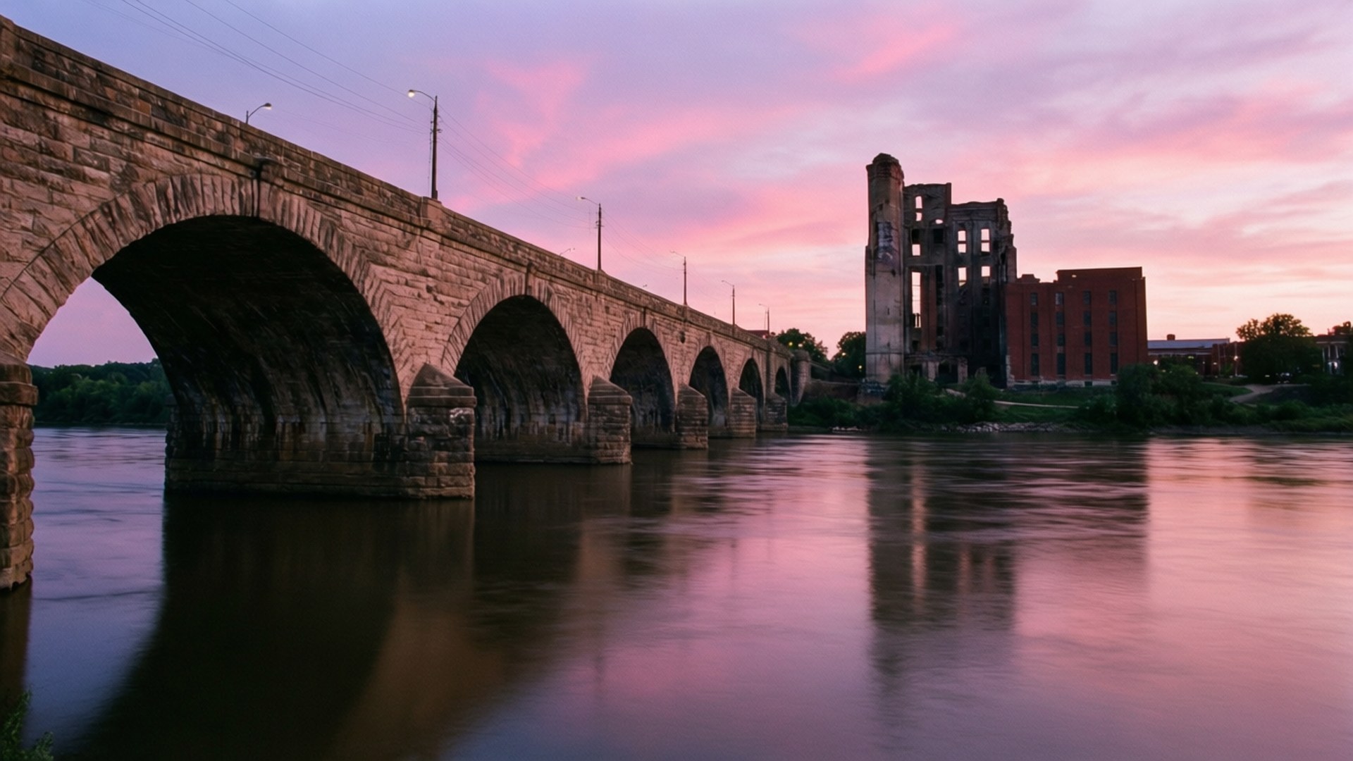 Stone Arch Bridge Twilight 壁紙 — 一座石拱橋橫跨平靜的河流，在充滿活力的粉紅色和紫色日落天空下，背景是一座廢棄的工廠。