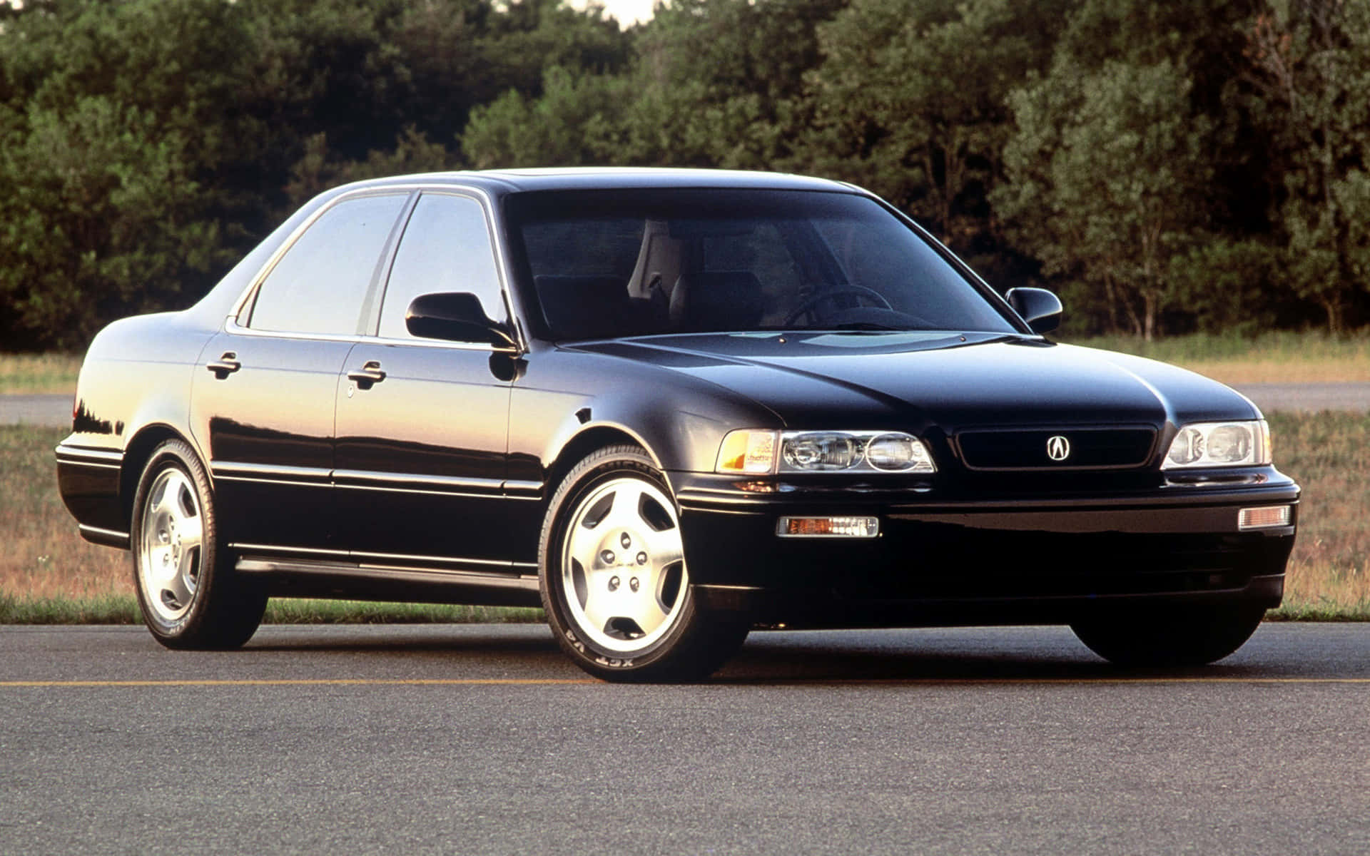 Acura Vigor 桌布 壁紙 — 一輛時尚的黑色Acura轎車在黃金光線下閃耀，行駛在兩旁茂密綠樹的寧靜道路上。