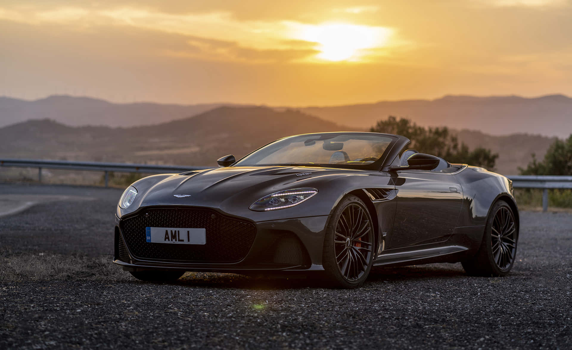 Aston Martin Vantage 桌布 壁紙 — 一架時尚的黑色Aston Martin DB11 Volante敞篷車停泊在碎石路上，背景是日落時分的山脈。