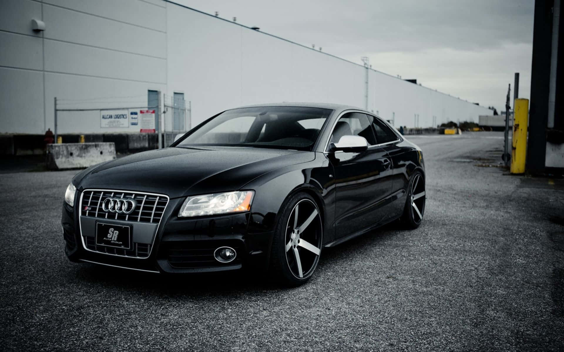 Audi S5 桌布 壁紙 — 一輛時尚的黑色奧迪轎跑車停在瀝青路上，前方是大型工業建築，天空多雲。