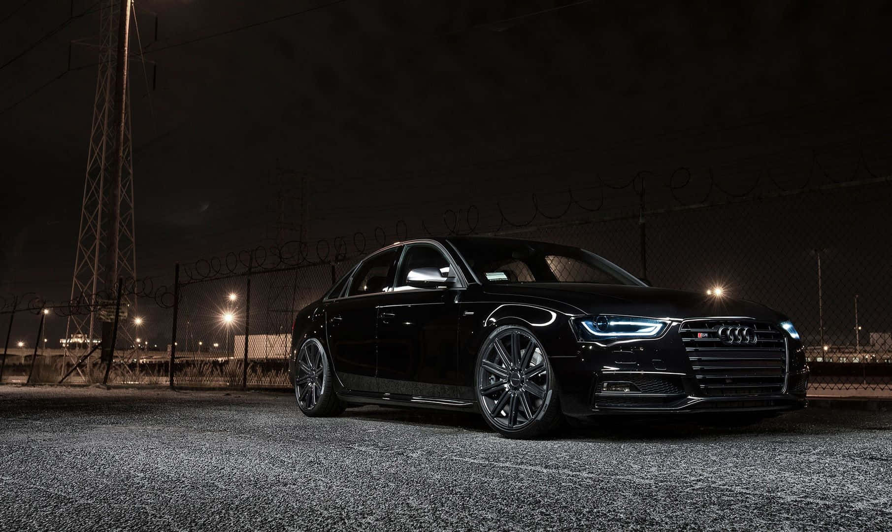 Audi A6 桌布 壁紙 — 一輛時尚的黑色奧迪轎車在夜燈下閃閃發光，停泊在帶有鐵絲網的鏈條圍欄旁。