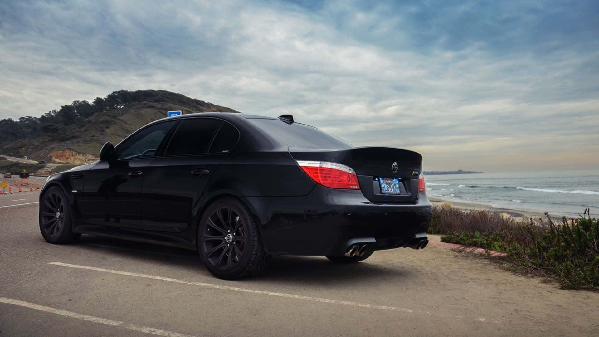 Bmw M5 桌布 壁紙 — 一輛時尚的黑色BMW轎車停泊在沿海公路上，海浪在附近拍打，天空多雲。