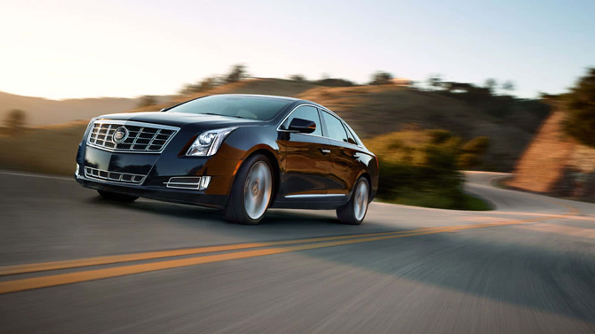 Cadillac Xts 桌布 壁紙 — 一輛時尚的黑色凱迪拉克轎車在日落時分沿著蜿蜒的道路疾馳，背景是模糊的山丘。