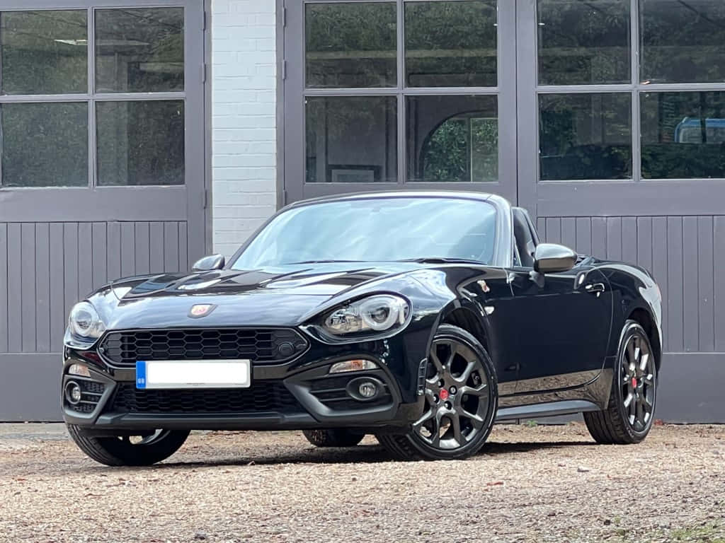 Fiat 124 Spider 桌布 壁紙 — 一架時尚黑色飛雅特124 Spider敞篷車停放在現代灰色車庫前的碎石地上，車庫有大型窗戶。