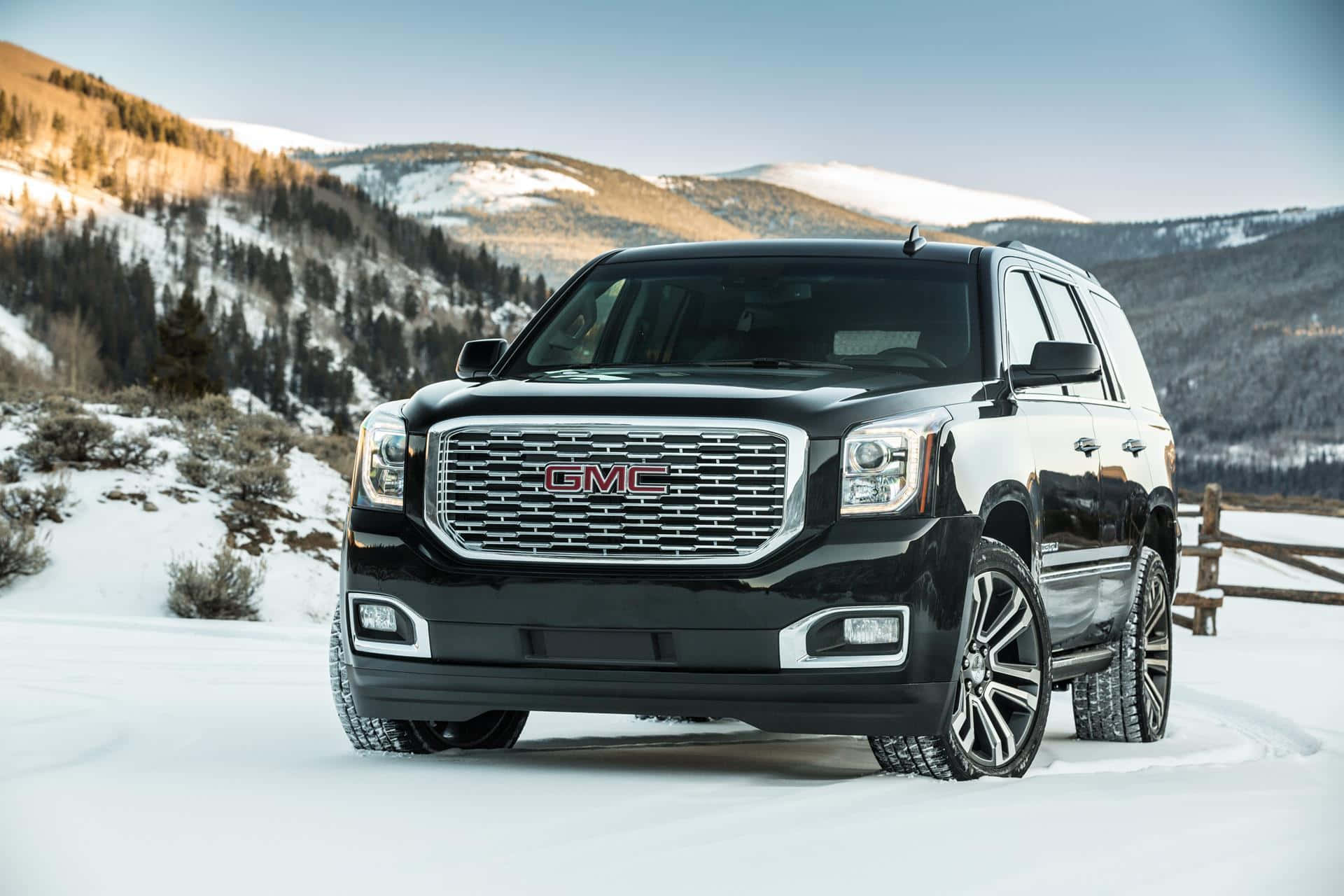 Gmc Yukon 桌布 壁紙 — 一輛時尚的黑色GMC Yukon SUV停在積雪的山路上，背景是白雪覆蓋的山峰和常青樹。