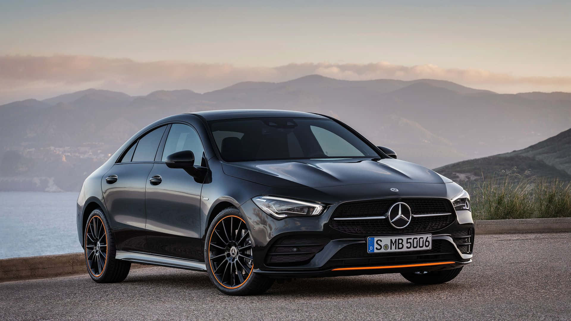 Mercedes Benz Cla-class 桌布 壁紙 — 一輛時尚的黑色賓士CLA轎車，帶有橙色點綴，停泊在日落時分俯瞰山巒和海洋的海岸公路上。