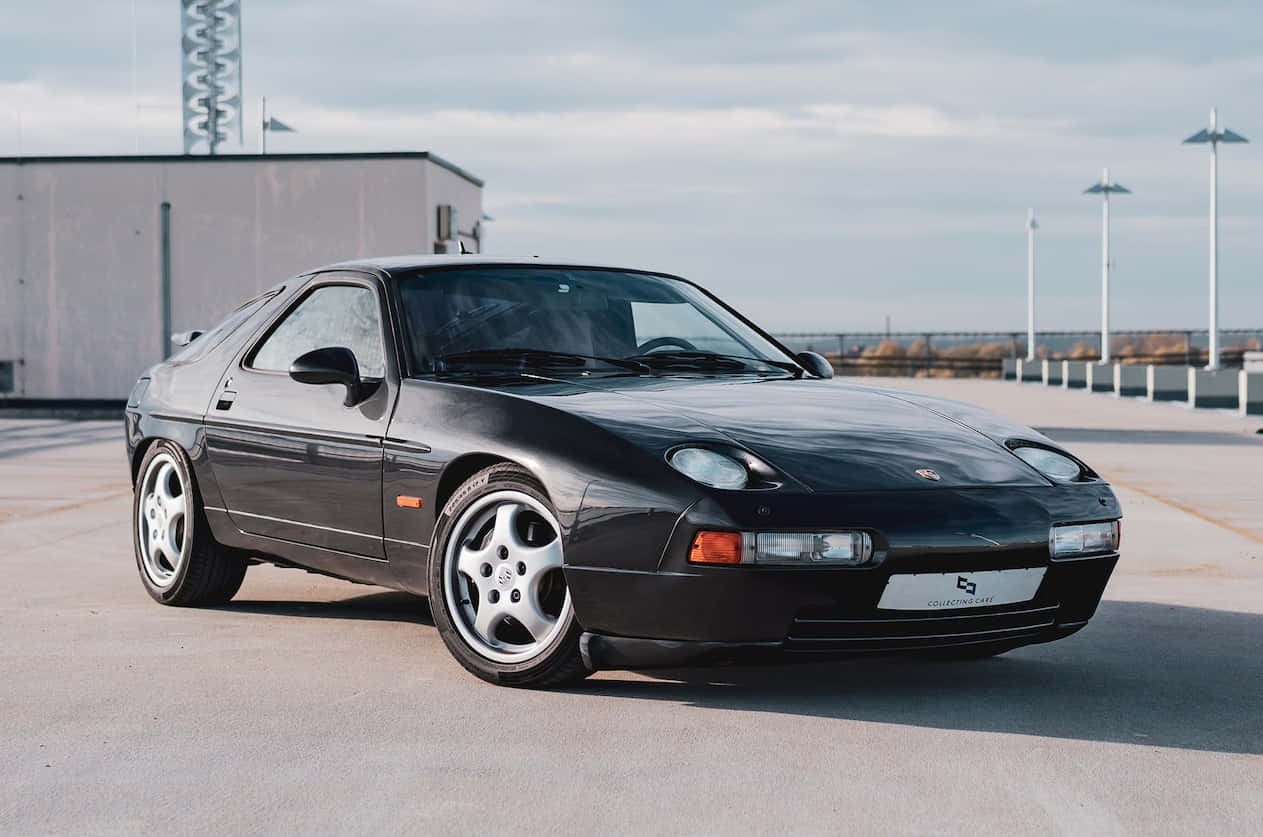 Porsche 928 桌布 壁紙 — 一輛時尚的黑色保時捷928跑車停放在混凝土停車場，天空多雲。